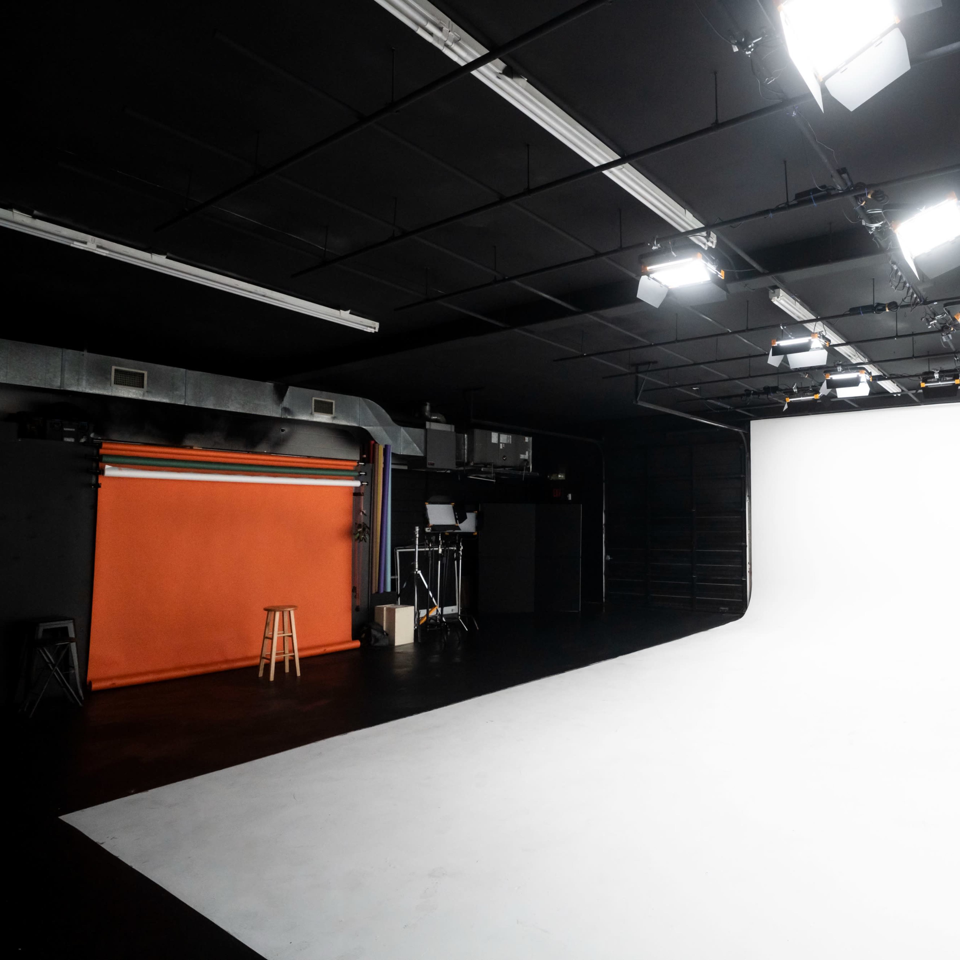 A photography studio features an orange backdrop, a stool, and multiple overhead lights illuminating a white backdrop area.