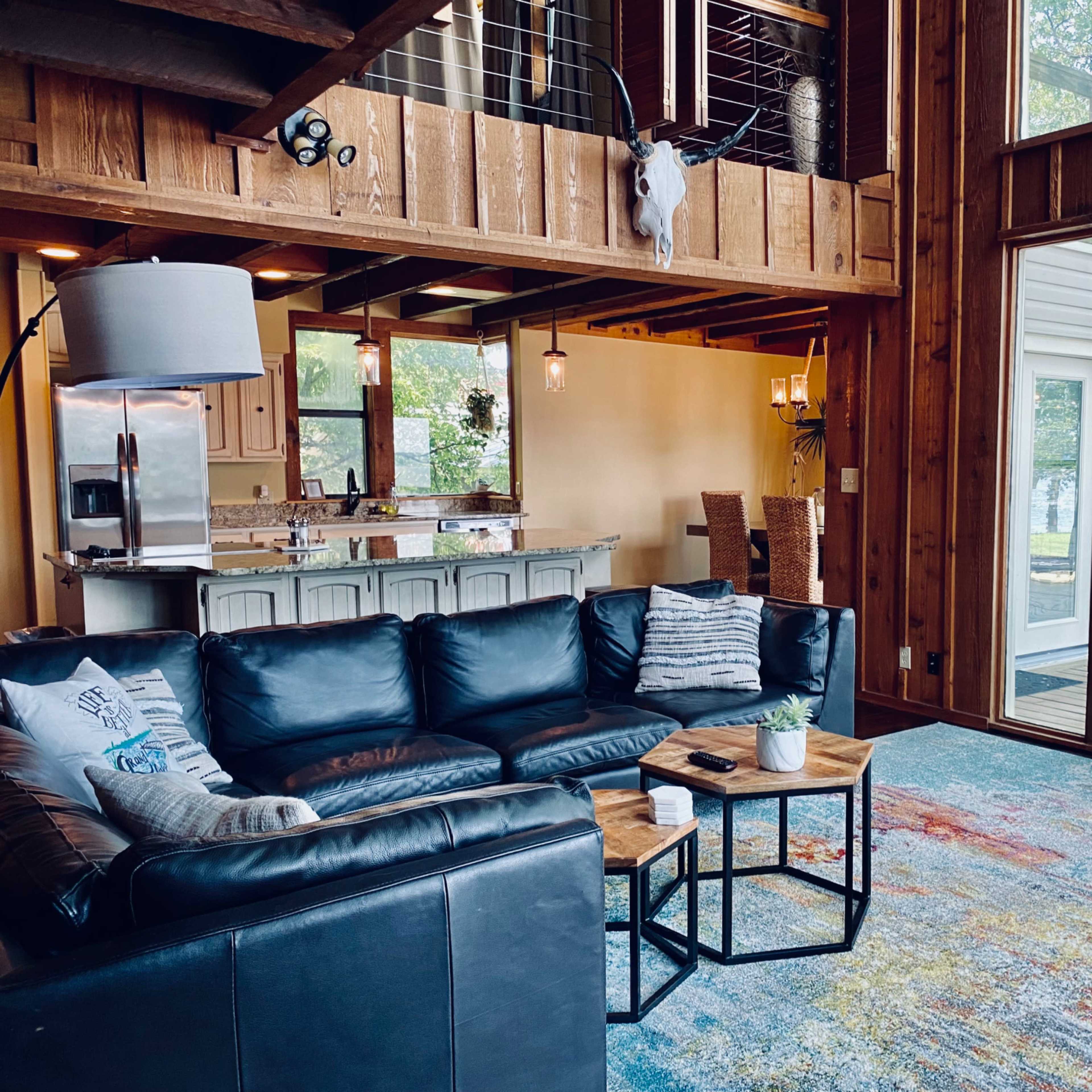 A spacious living room with a large black sectional sofa, wooden accents, and a view of the outdoors through large windows.