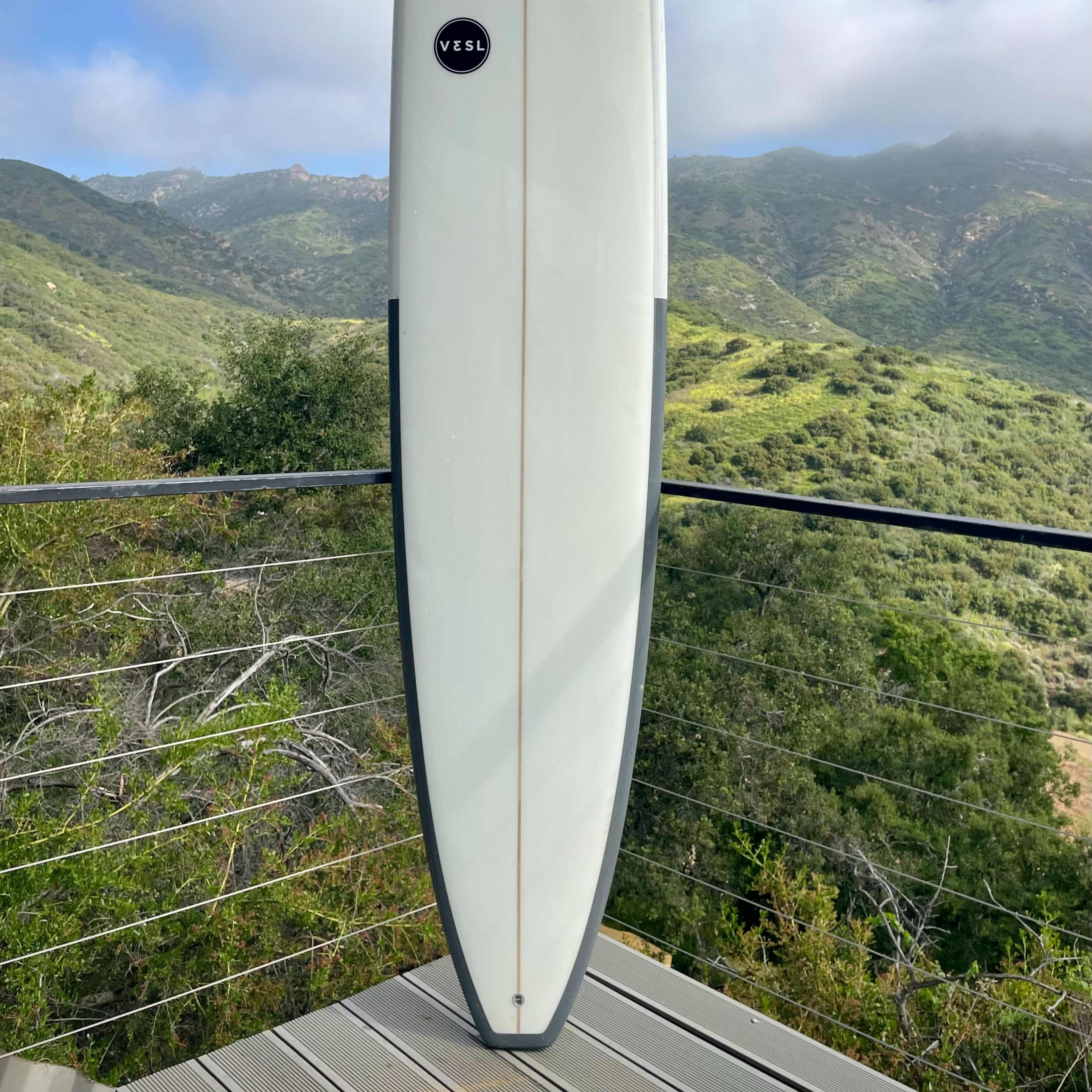 A longboard surfboard stands upright on a balcony with green hills and cloudy skies in the background.