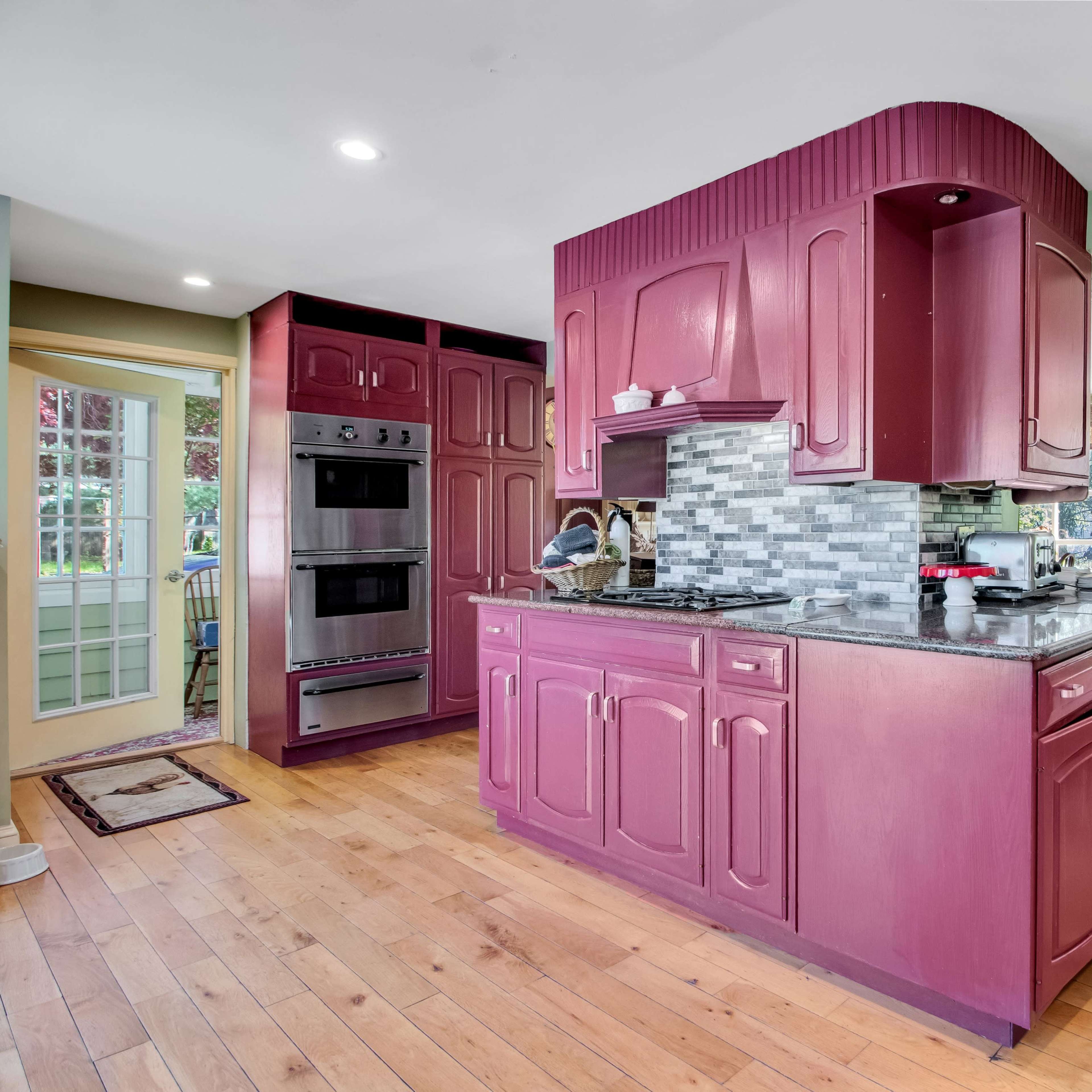 A kitchen with maroon cabinets and a granite countertop, featuring modern appliances and an adjoining dining area with wooden furniture.