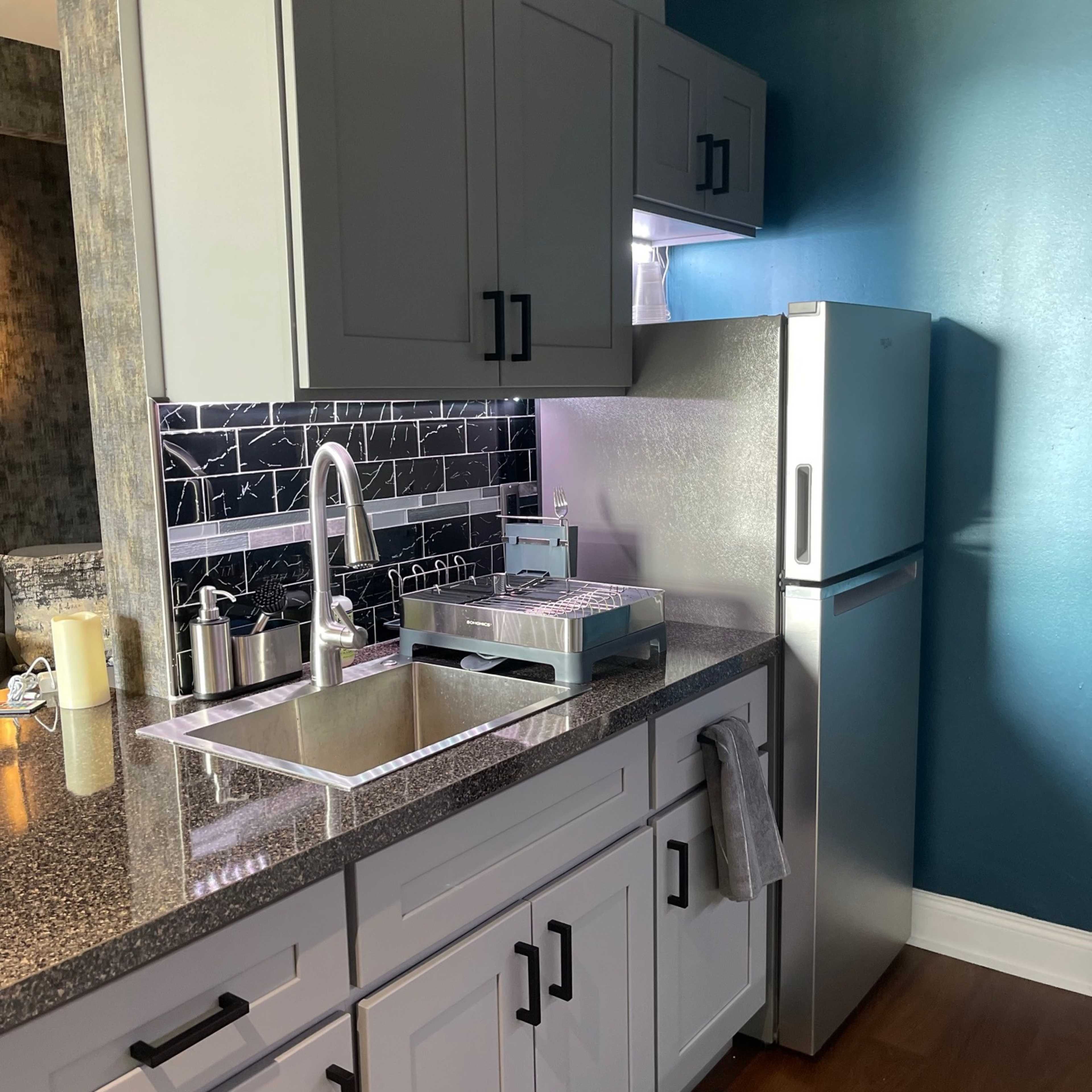 A modern kitchen with gray cabinets, a stainless steel refrigerator, a granite countertop, and a sink with a faucet.
