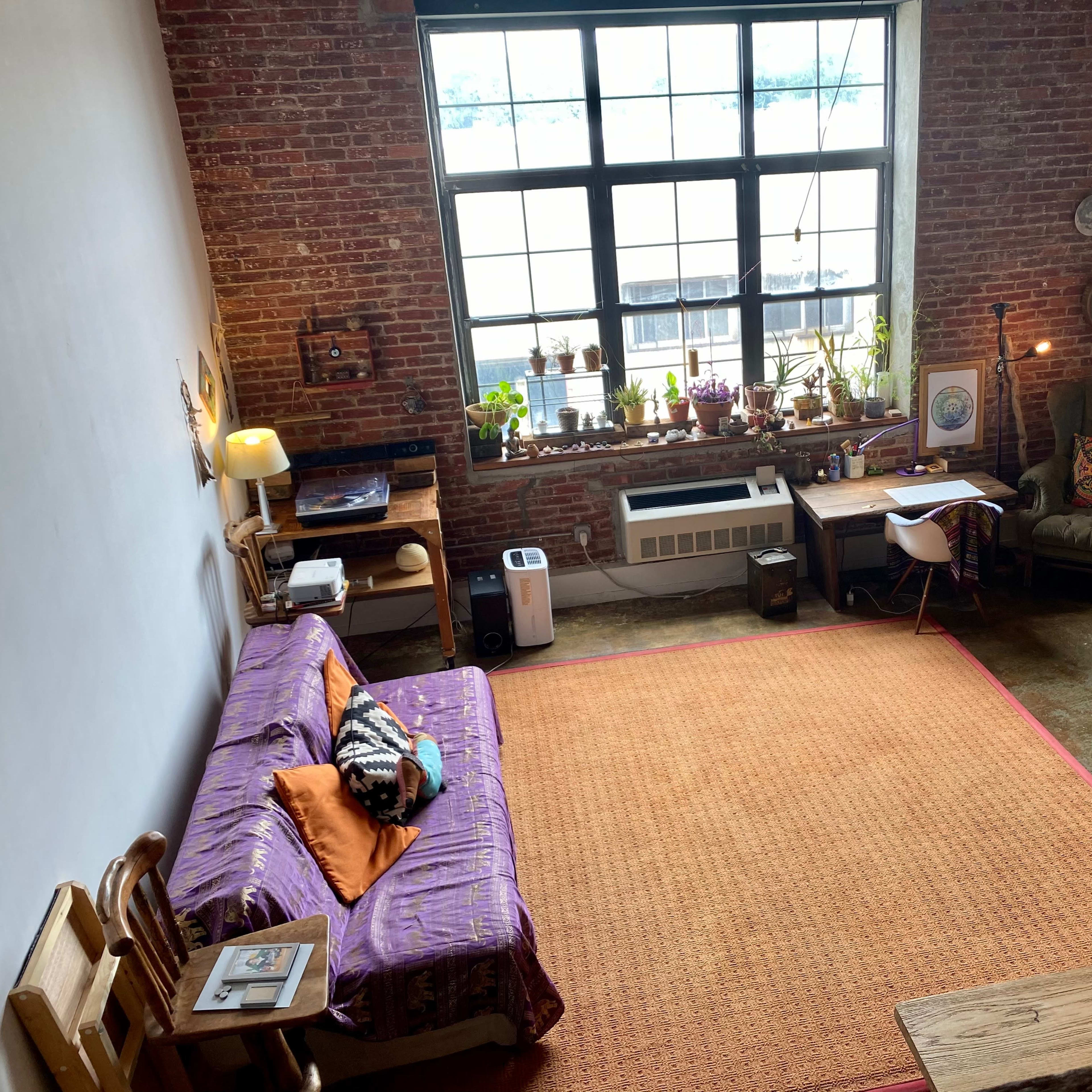 A bright, spacious living area featuring exposed brick walls, a large window, a sofa with colorful cushions, a wooden desk, and various plants.