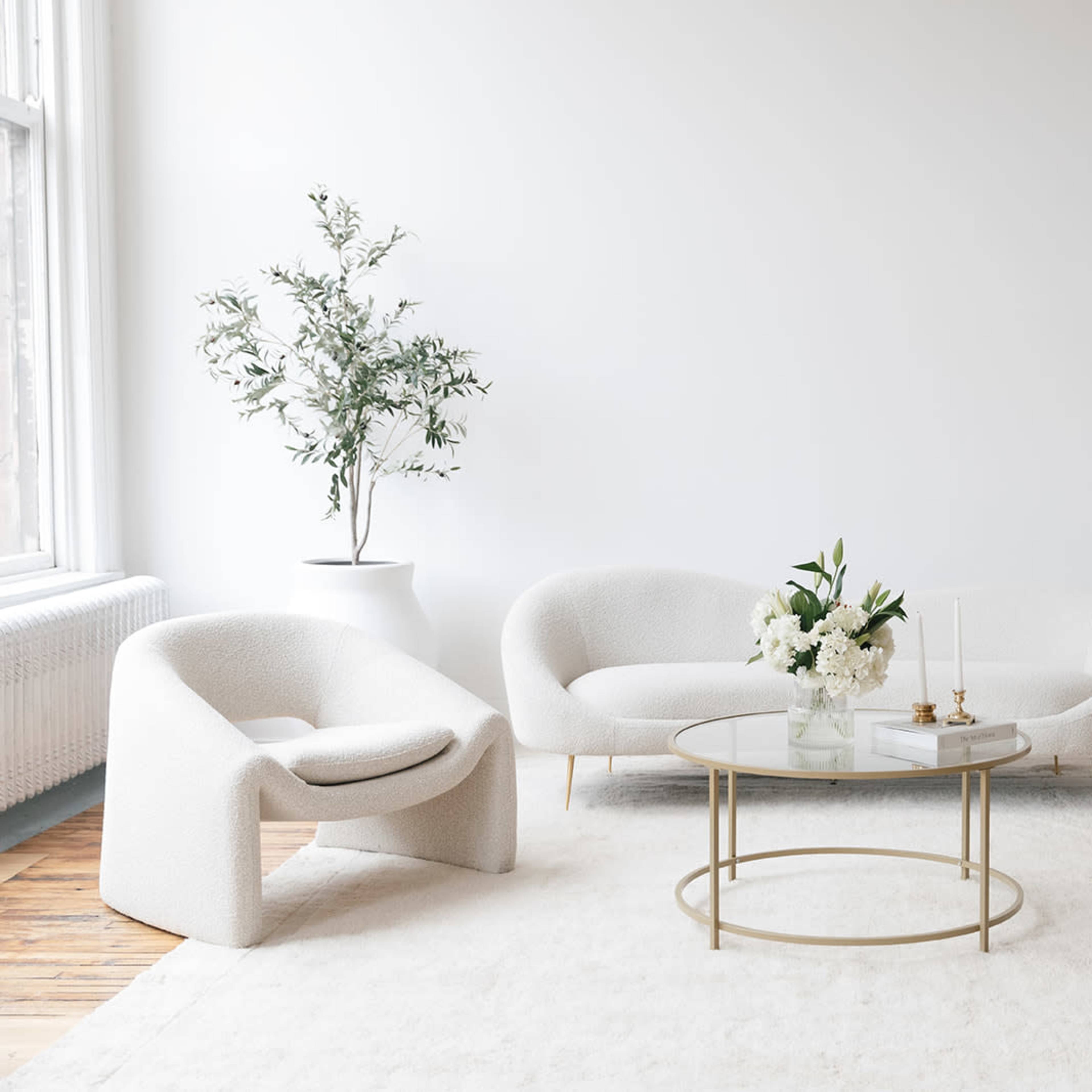 A bright, minimalistic living room featuring two white, curved sofas and two matching armchairs arranged around a glass coffee table with a floral centerpiece.