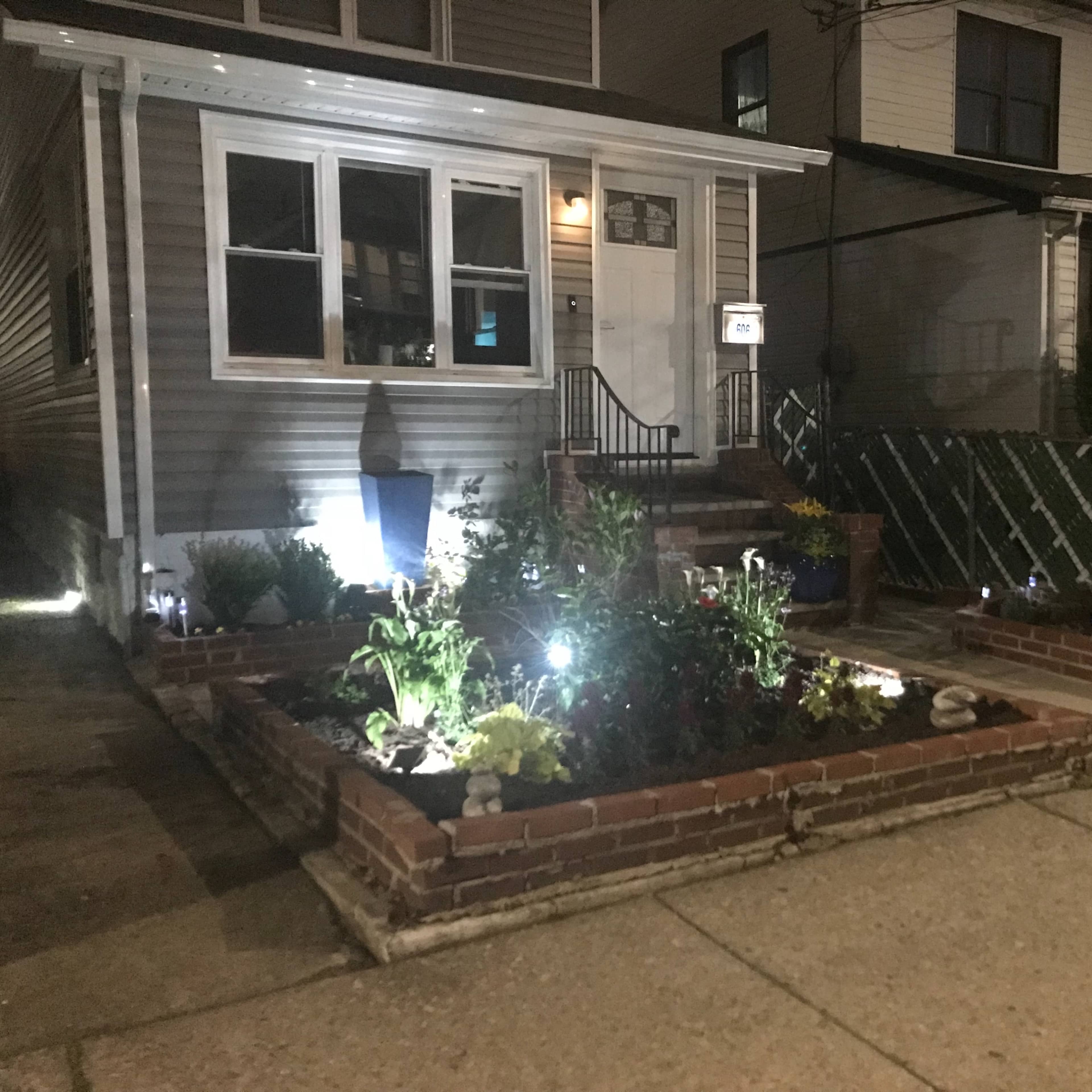 A house with a well-lit front garden featuring brick borders, plants, and decorative elements at night.