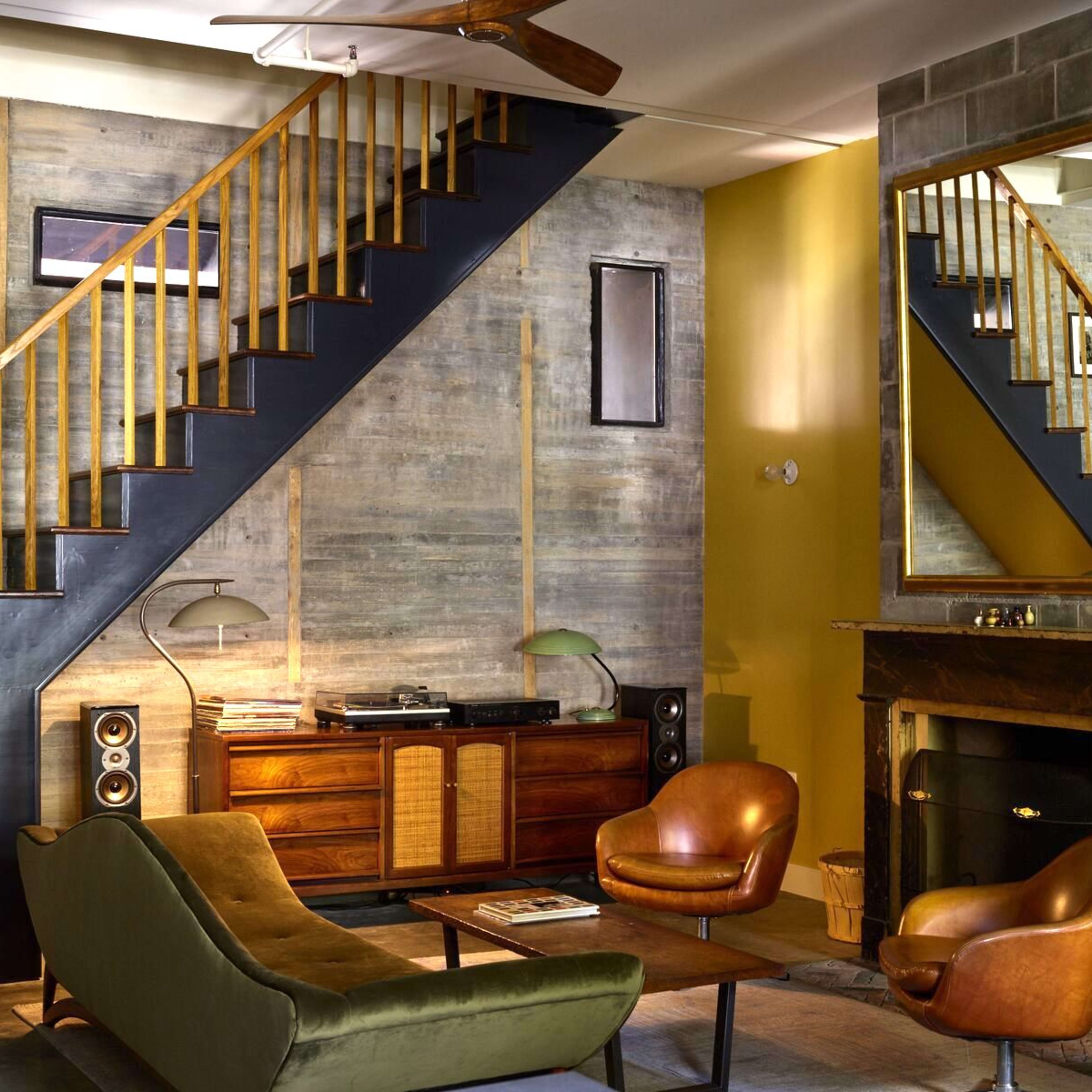 A modern living room with a green sofa, two brown armchairs, a wooden sideboard, a fireplace, and a staircase with a wooden railing against a textured wall.