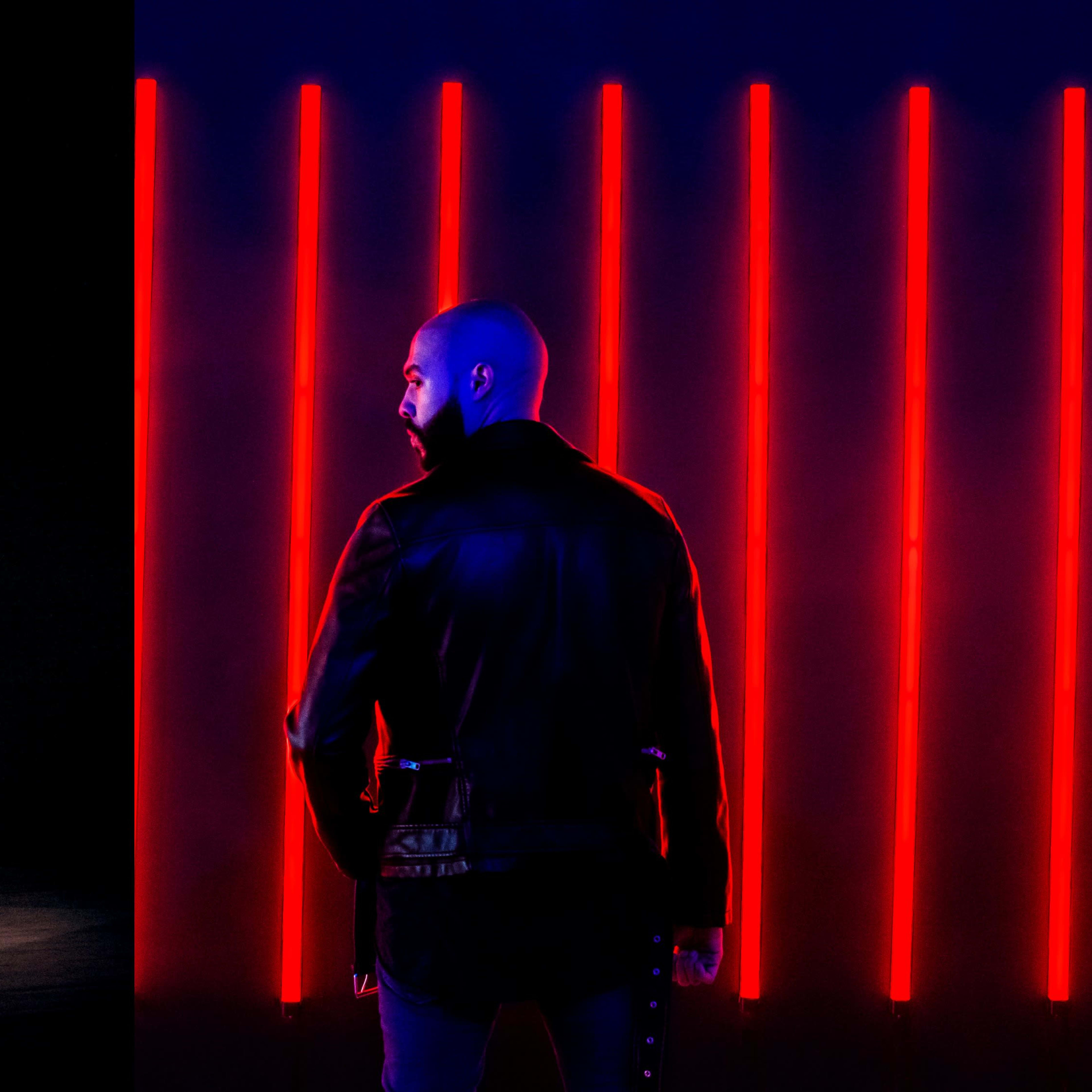 Two contrasting scenes: on the left, a person crouches in a spotlight with a glowing geometric outline, and on the right, another individual stands with their back to the viewer in front of vertical red lights.
