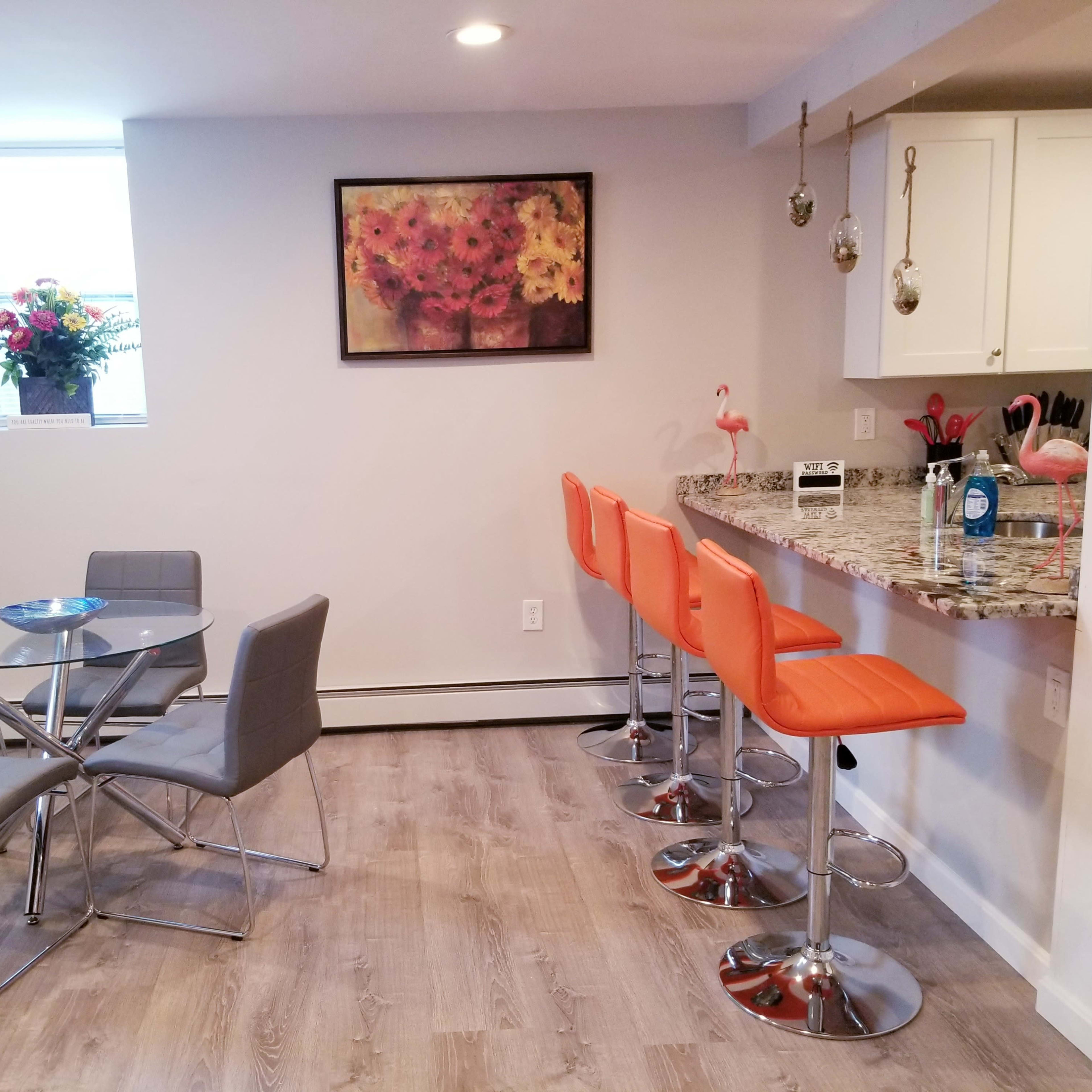 A modern kitchen and dining area featuring a circular glass table with gray chairs and a bar counter with orange stools.