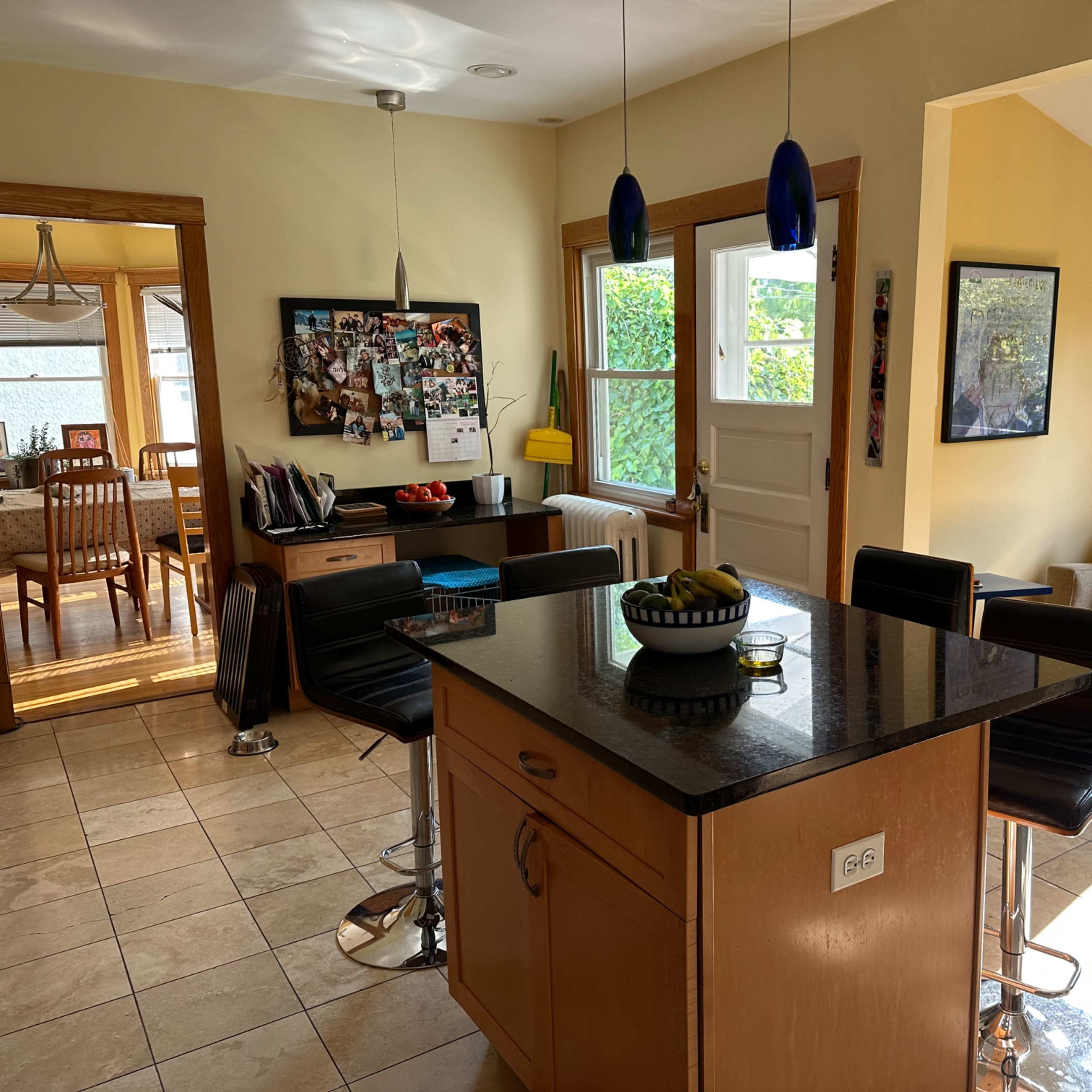 A kitchen with a central island, black bar stools, a dining area visible in the background, and a cozy seating area with a couch and a window.