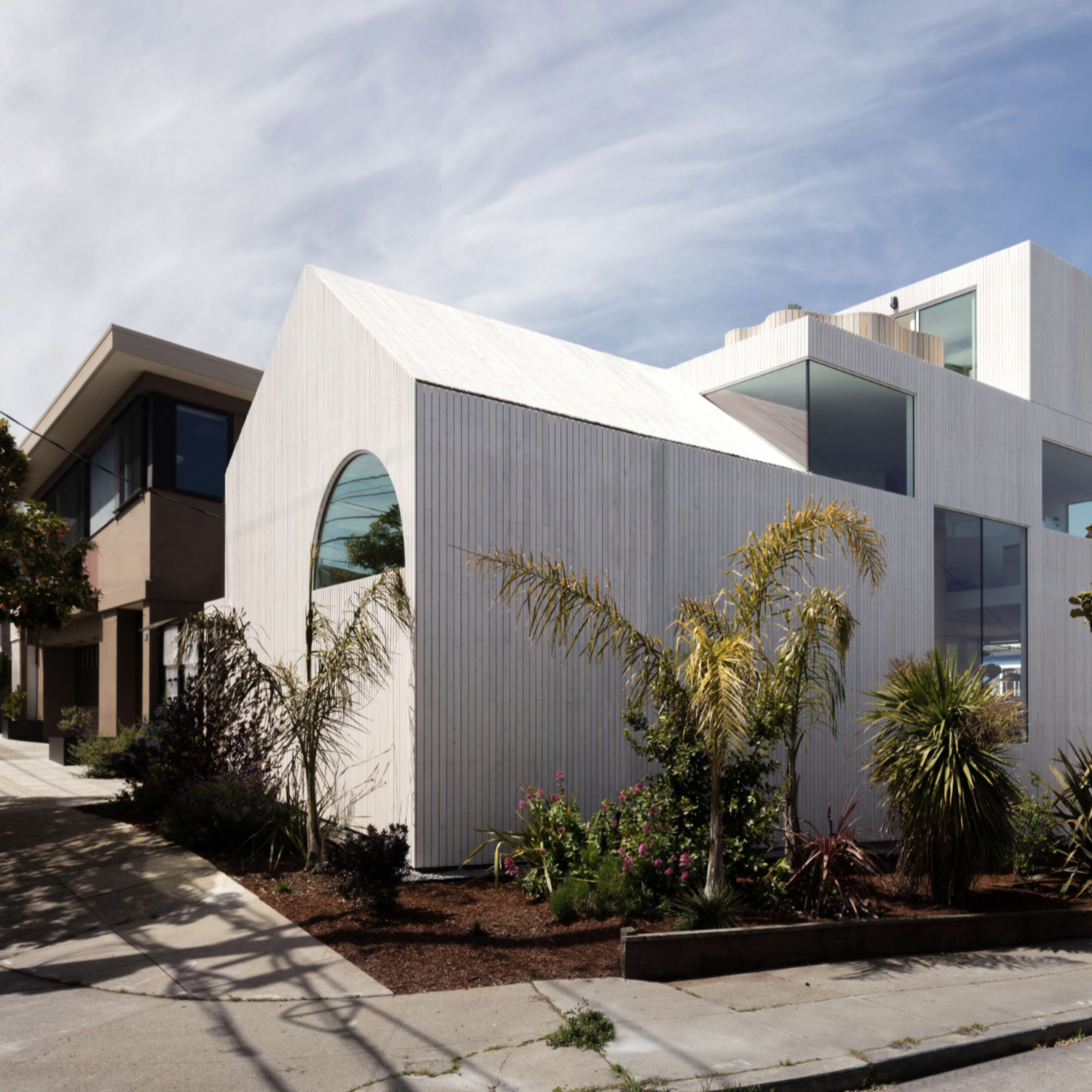A modern, angular house with a white wood exterior and large windows is located on a sloped street, surrounded by landscaped greenery and other residential buildings.