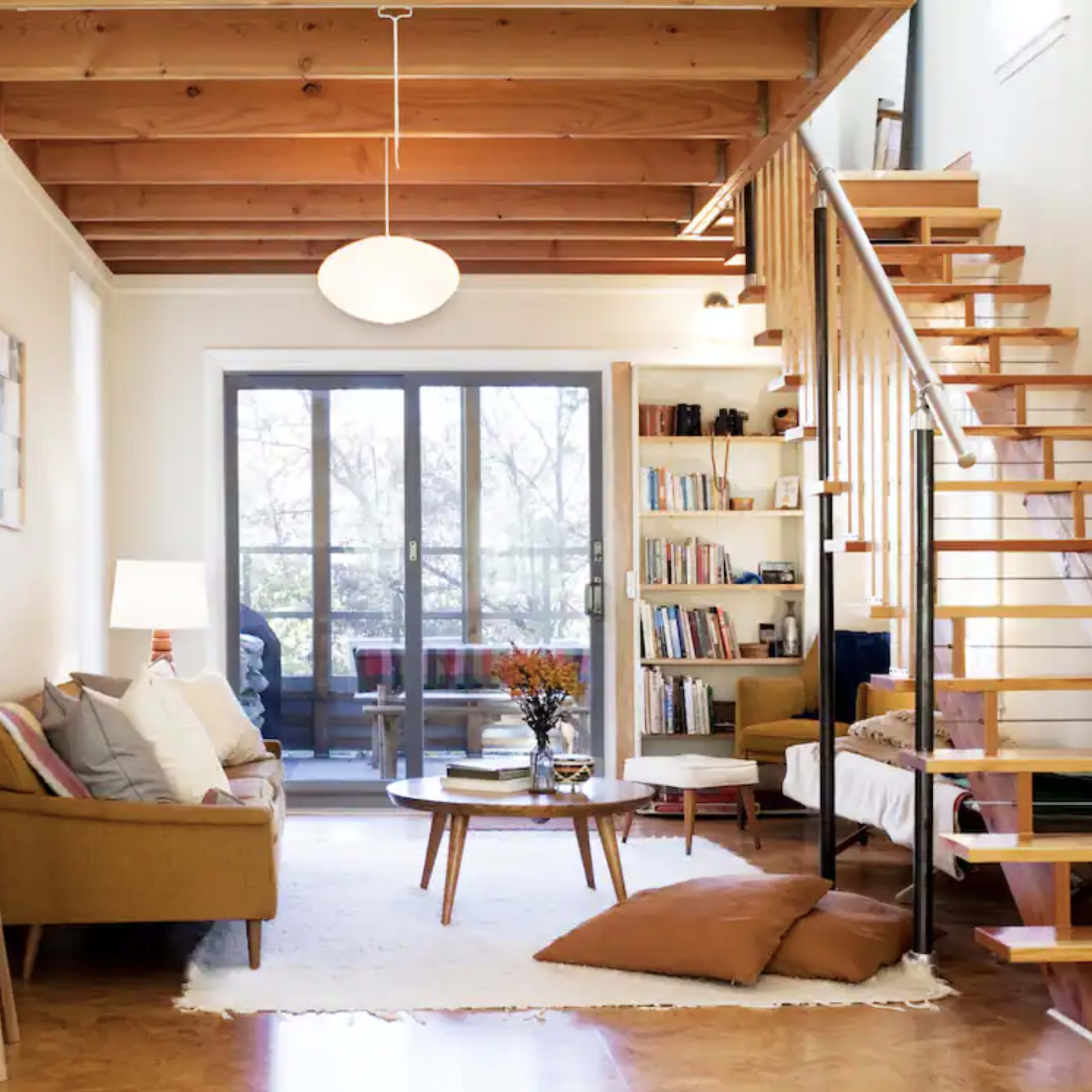 A modern living room with a wooden staircase, large windows, and a mix of seating arrangements including a sofa and armchairs.