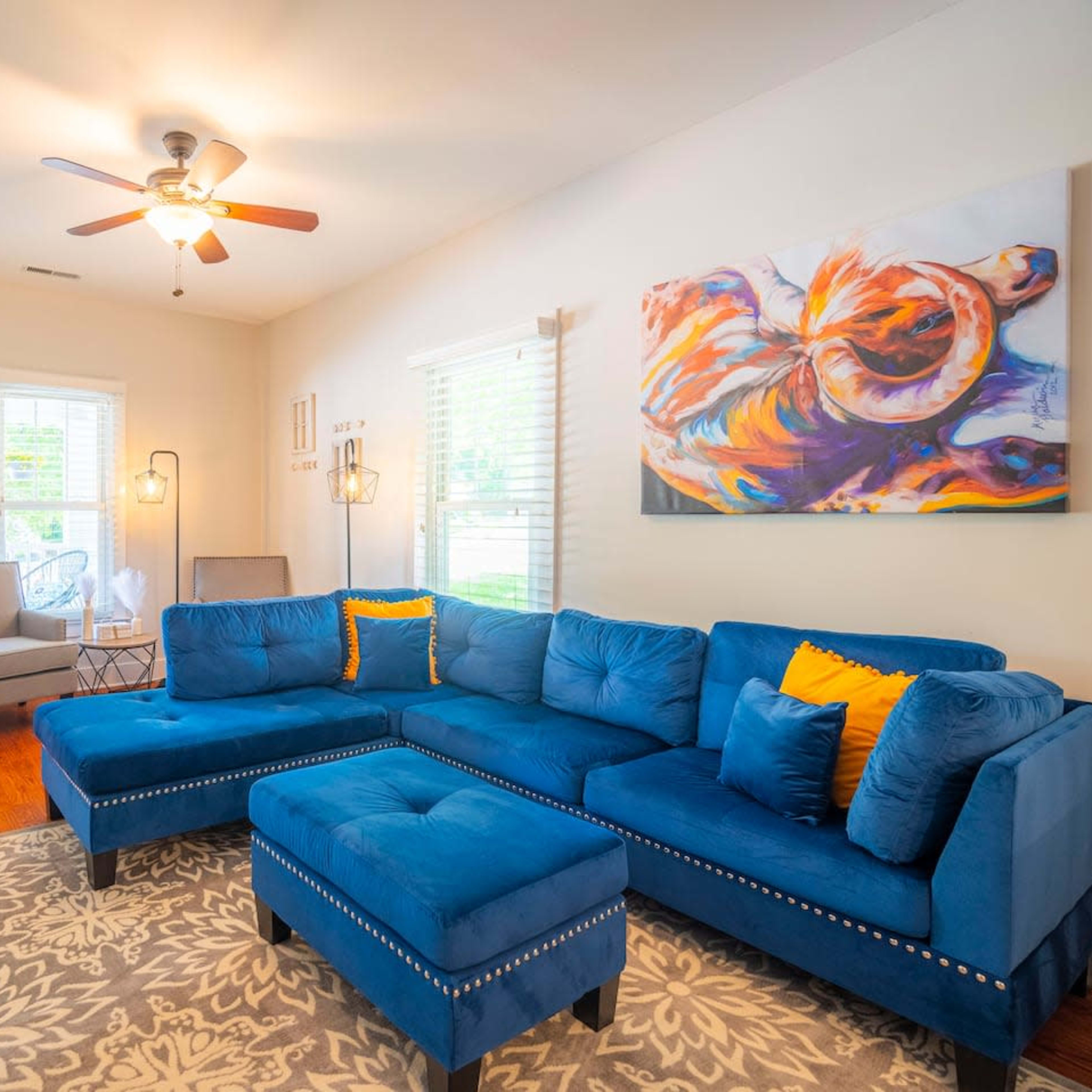 The living room features a blue sectional couch with decorative cushions, a ceiling fan, an area rug, and a colorful piece of wall art.