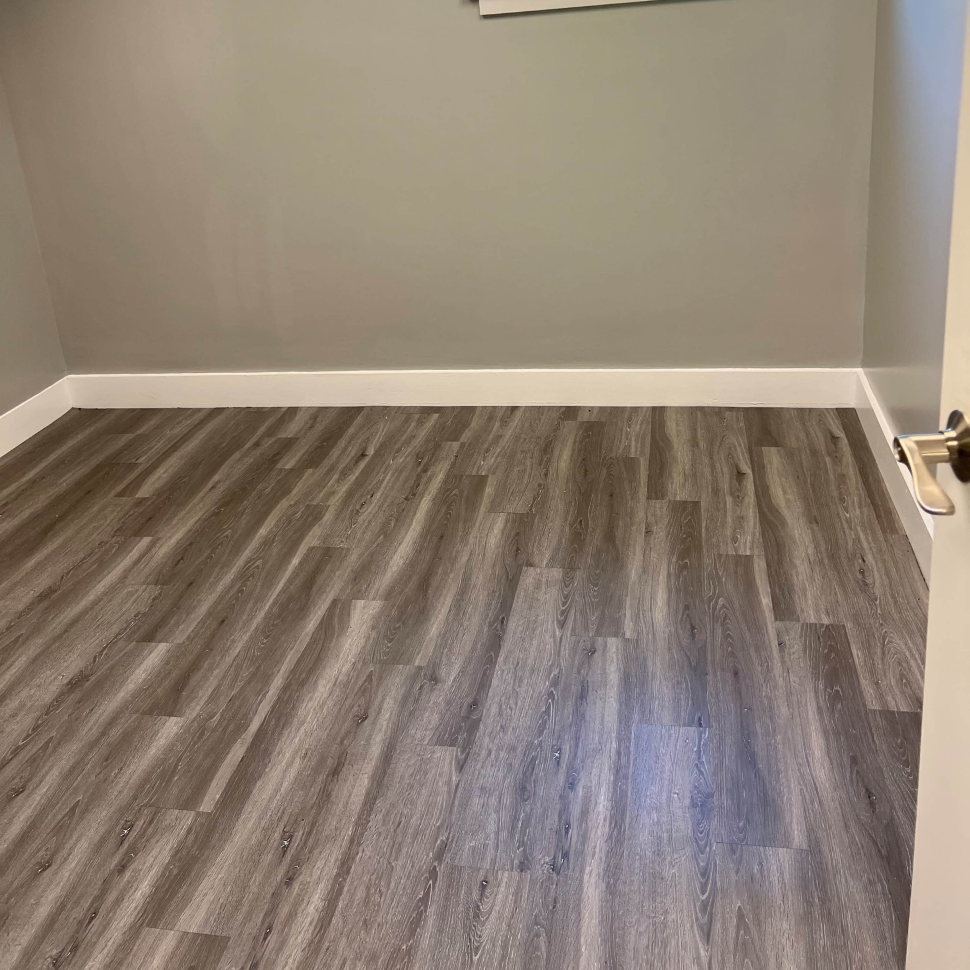 An empty room with gray walls, a window allowing natural light to enter, and dark wood-like flooring.