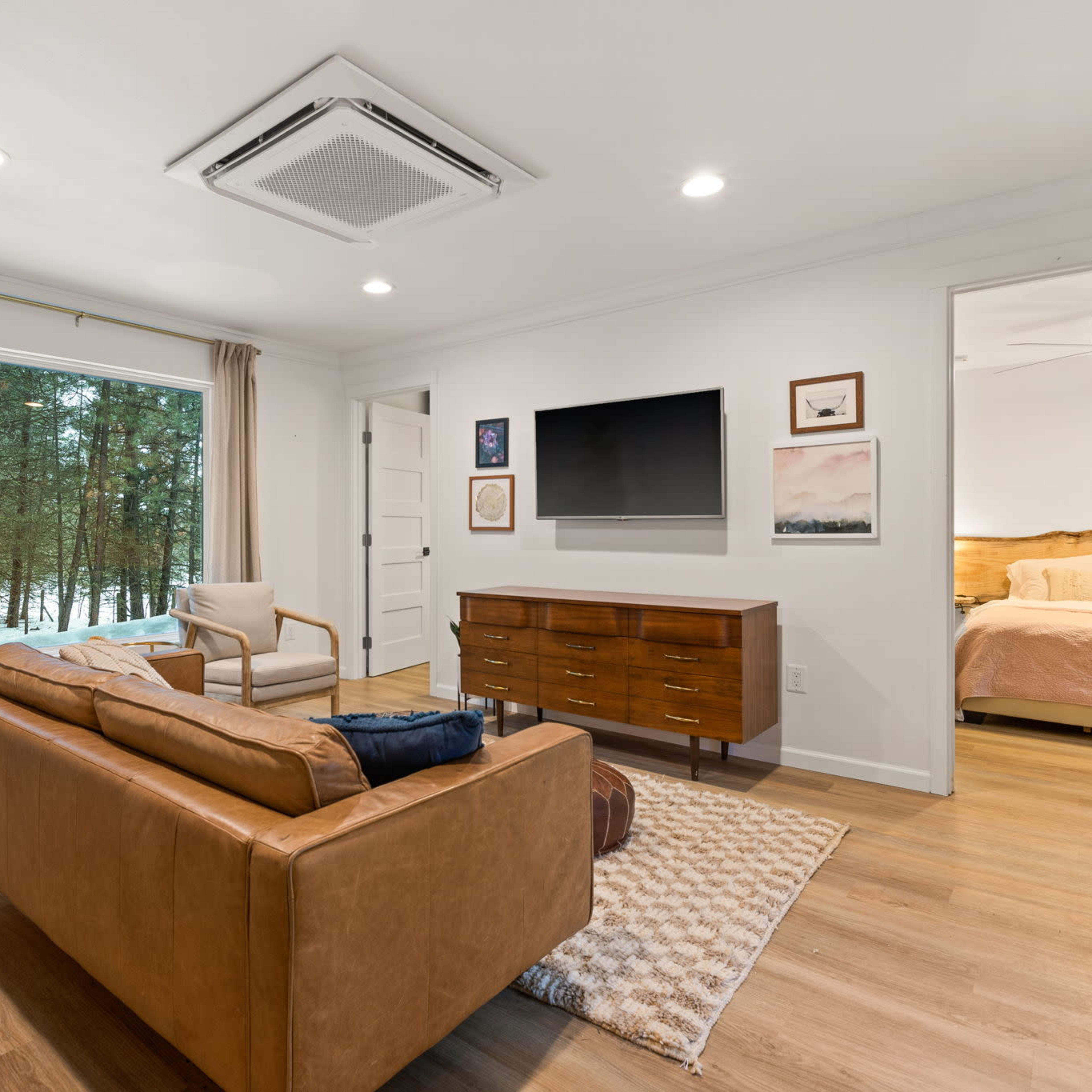 The living room features a leather sofa, a TV mounted on the wall, and a large window overlooking a forested area.
