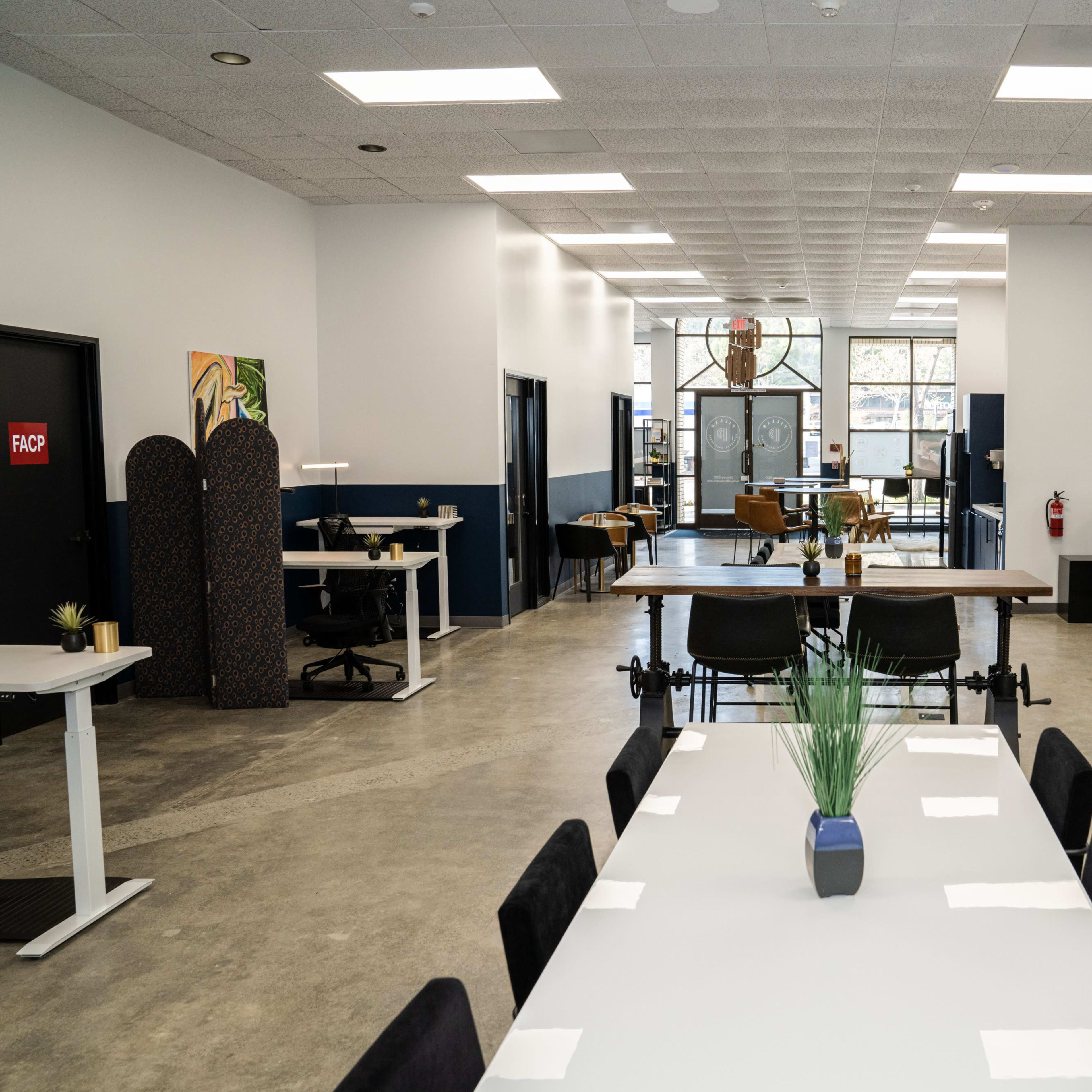 A modern office space with a large communal table, individual desks, and a glass-walled meeting room.