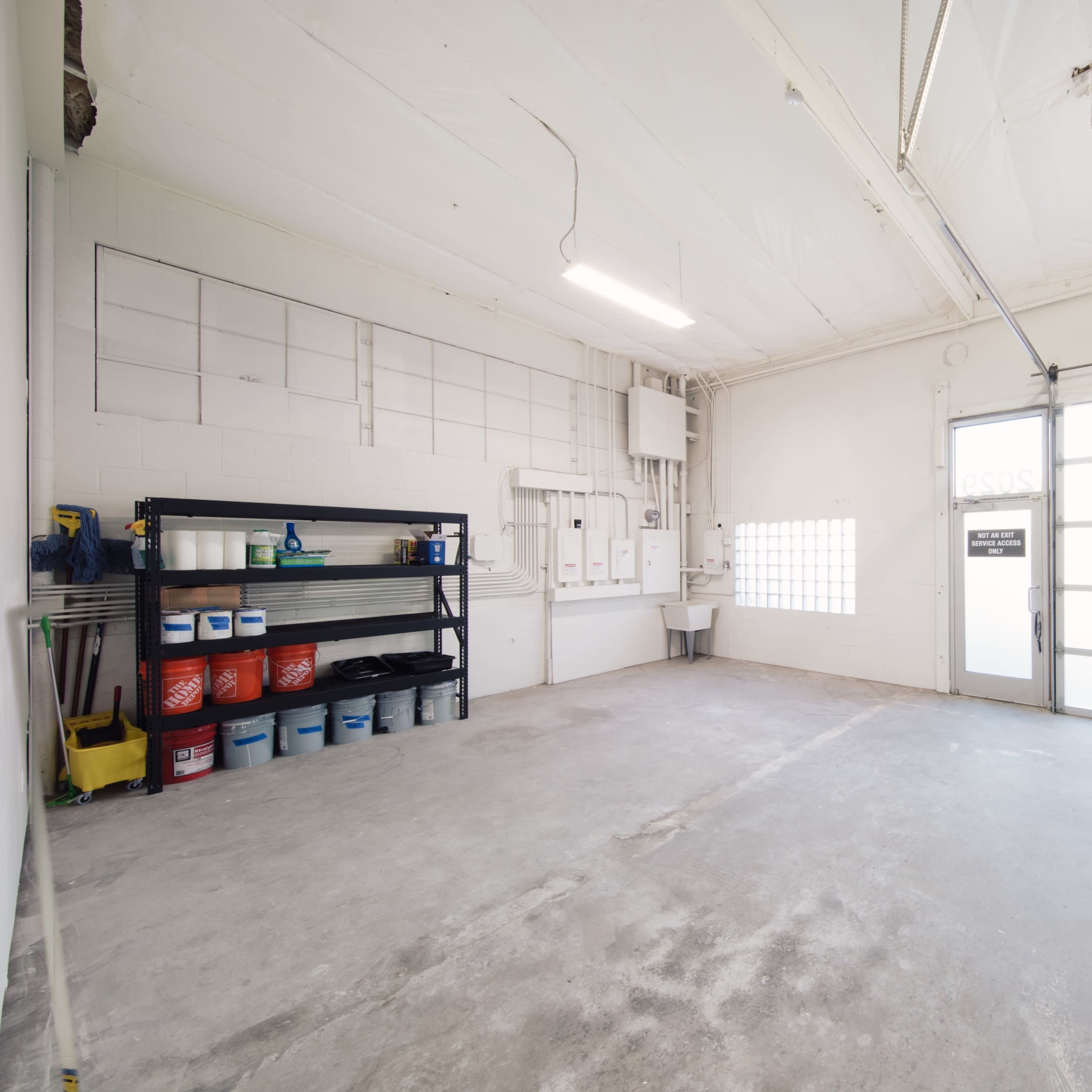 An empty garage with a concrete floor, a large roll-up door, and shelves filled with various storage containers.