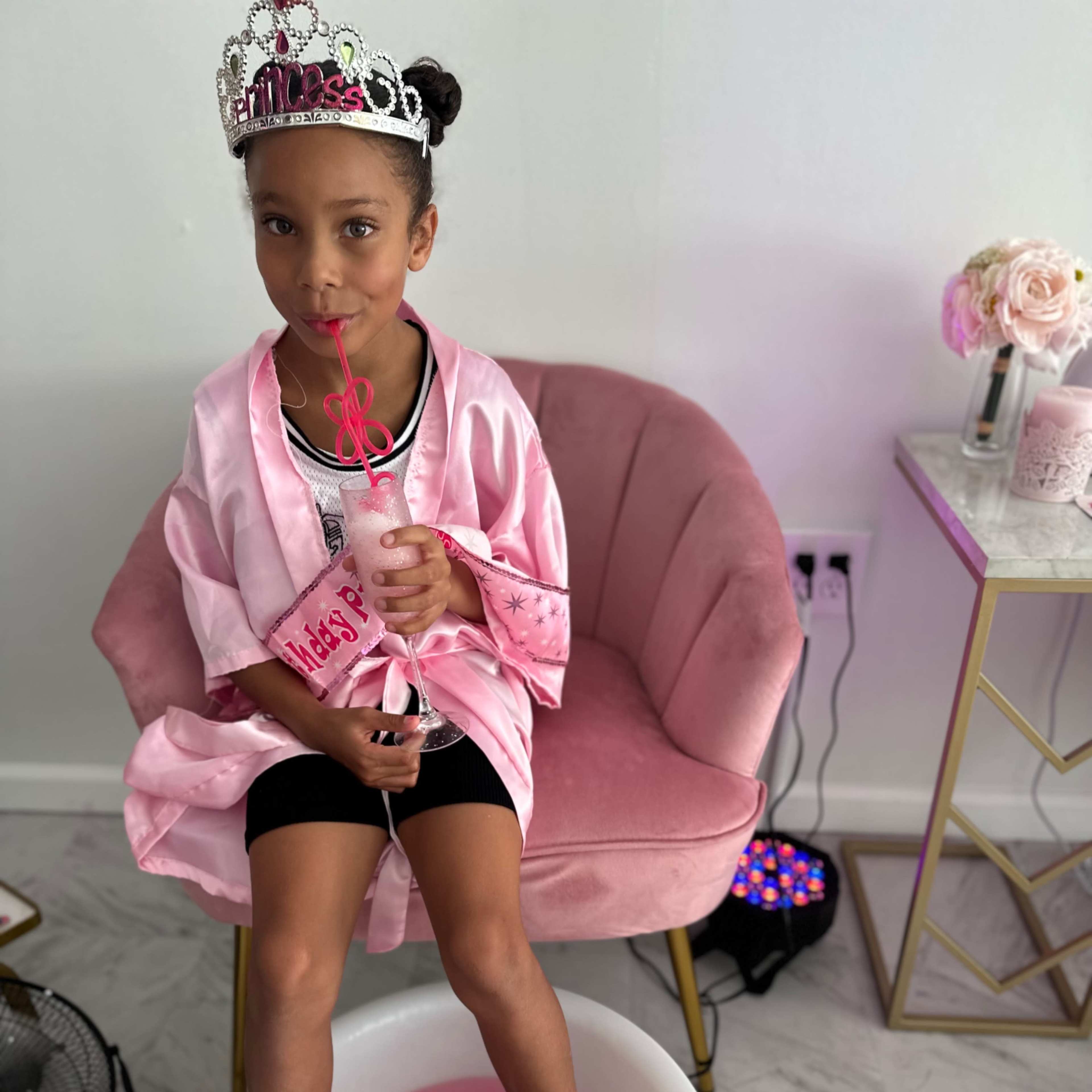 A child wearing a pink robe and a tiara sits on a pink chair with her feet in a bowl of pink water, holding a drink with a straw.