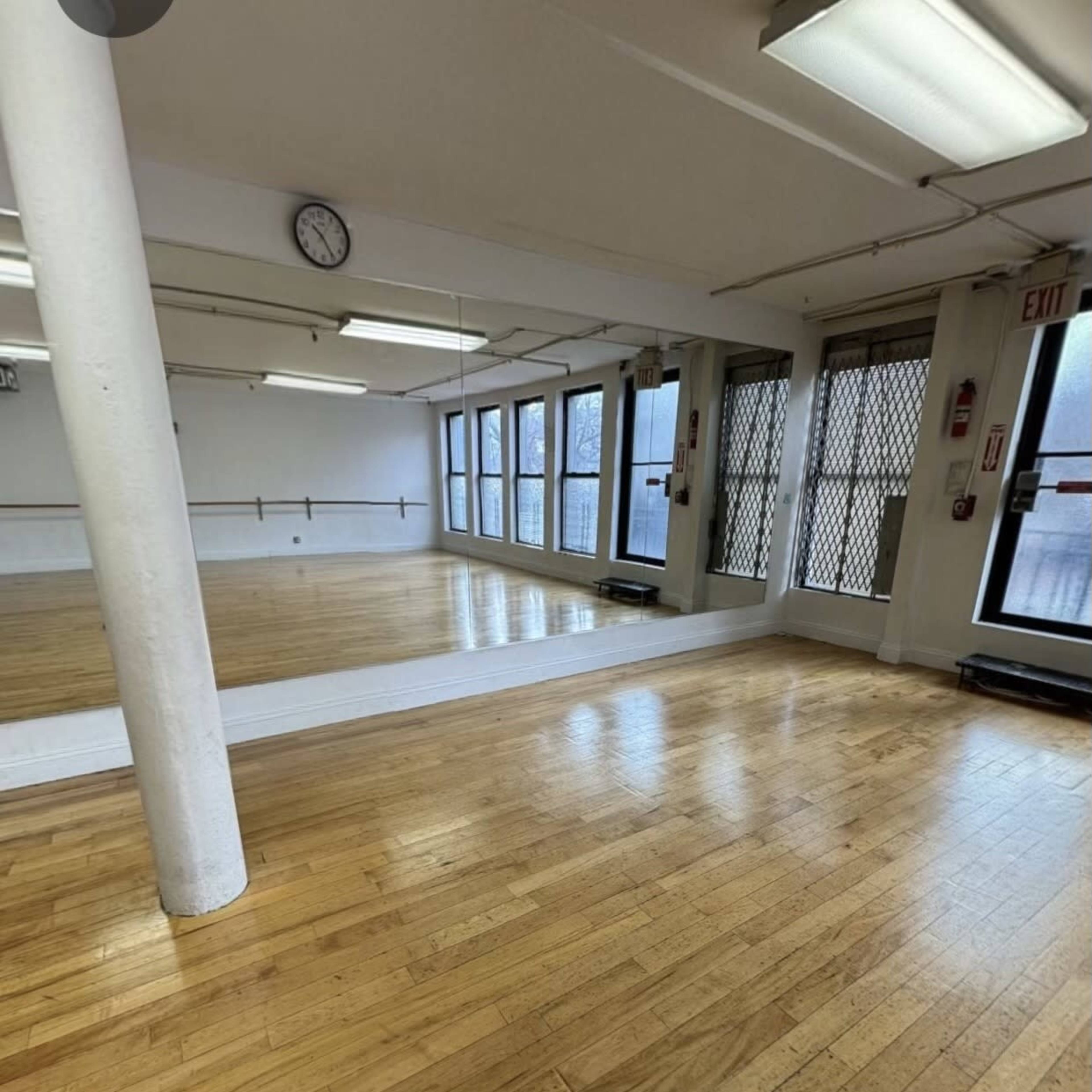 A spacious dance studio with wooden flooring, large windows, and a mirror along one wall.