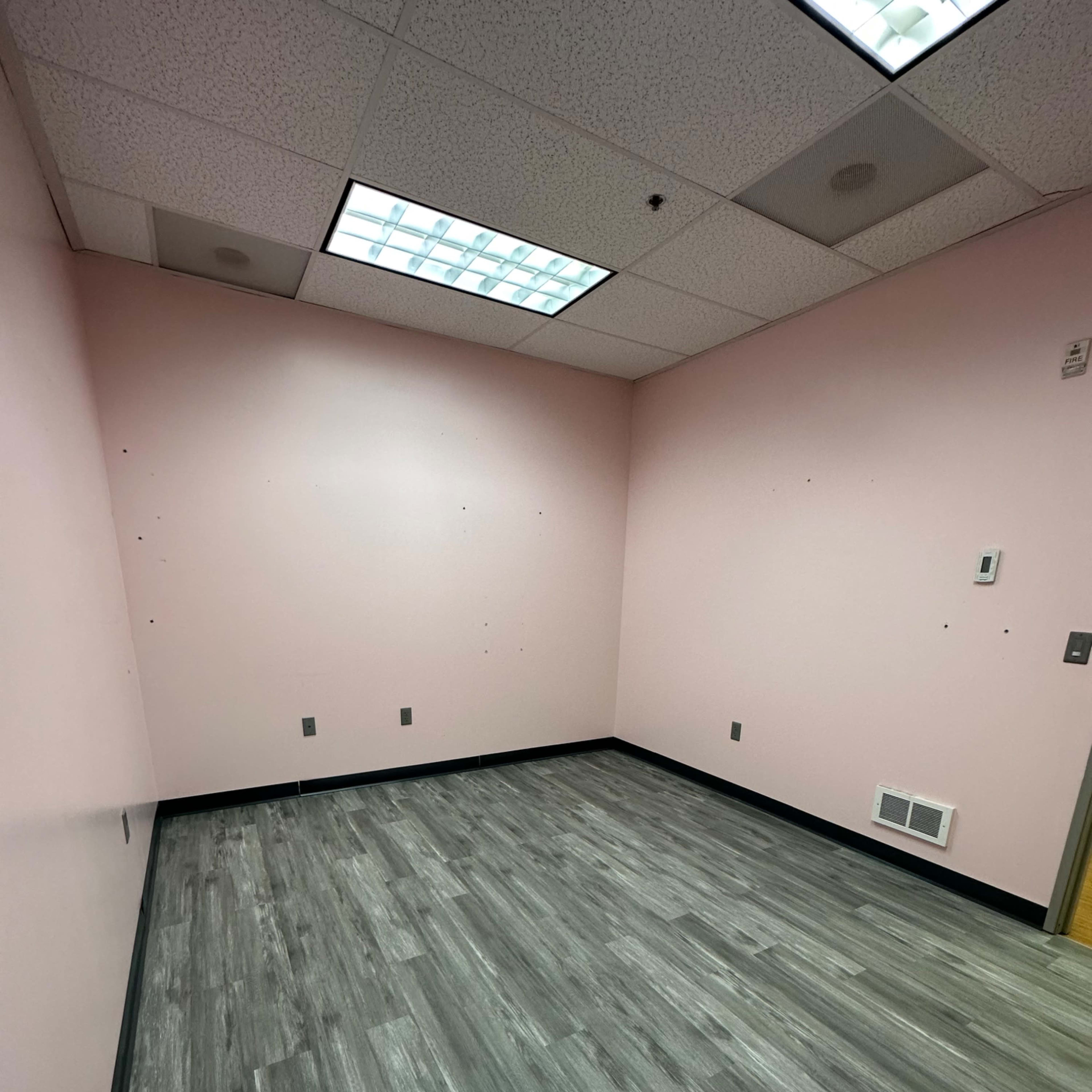 A vacant room with pink walls and gray laminate flooring, featuring a ceiling with fluorescent lights.