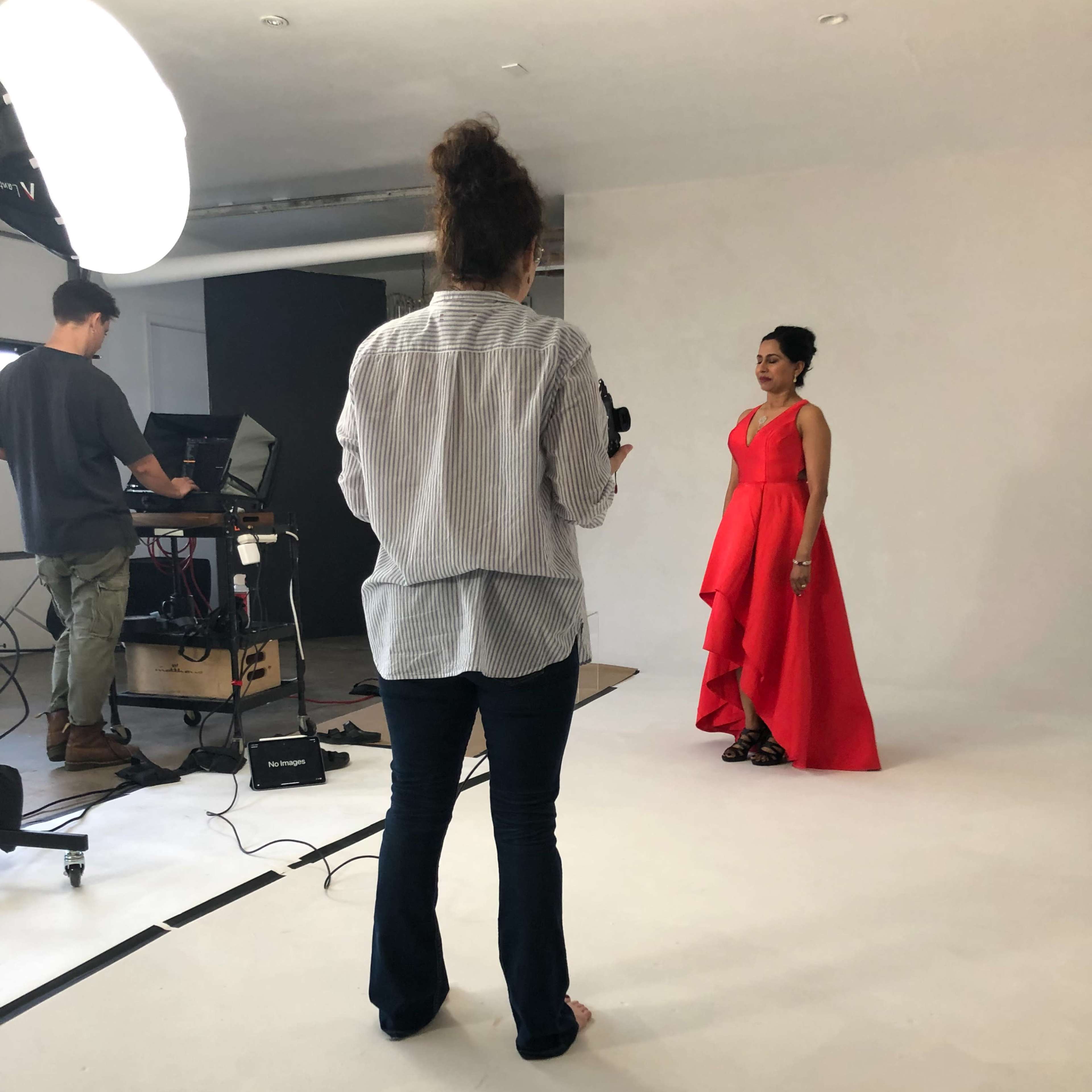 A woman in a red dress poses for a photo shoot while a photographer adjusts equipment in the background.