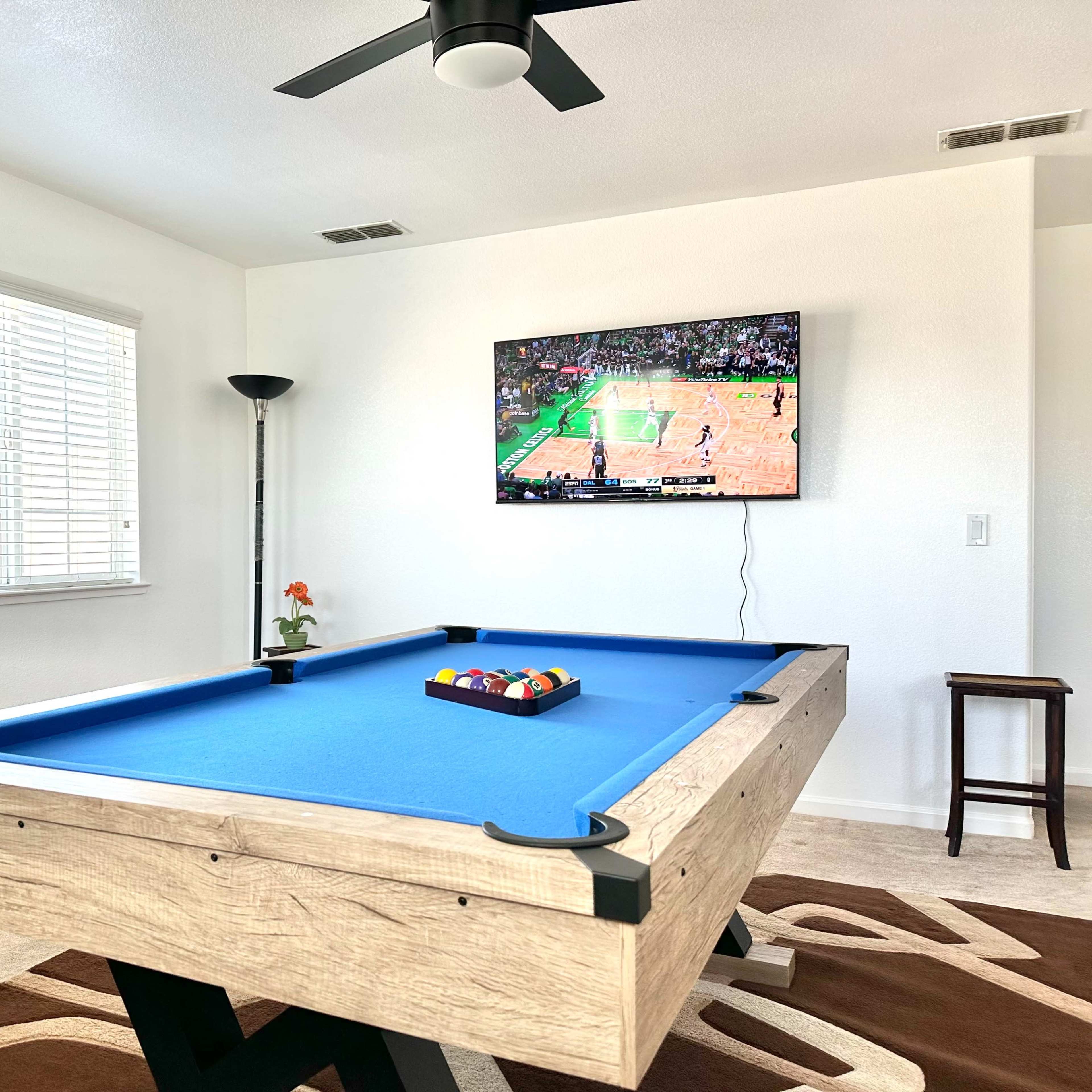 A game room with a blue pool table, a television displaying a basketball game, and a small flower in a pot on the table.