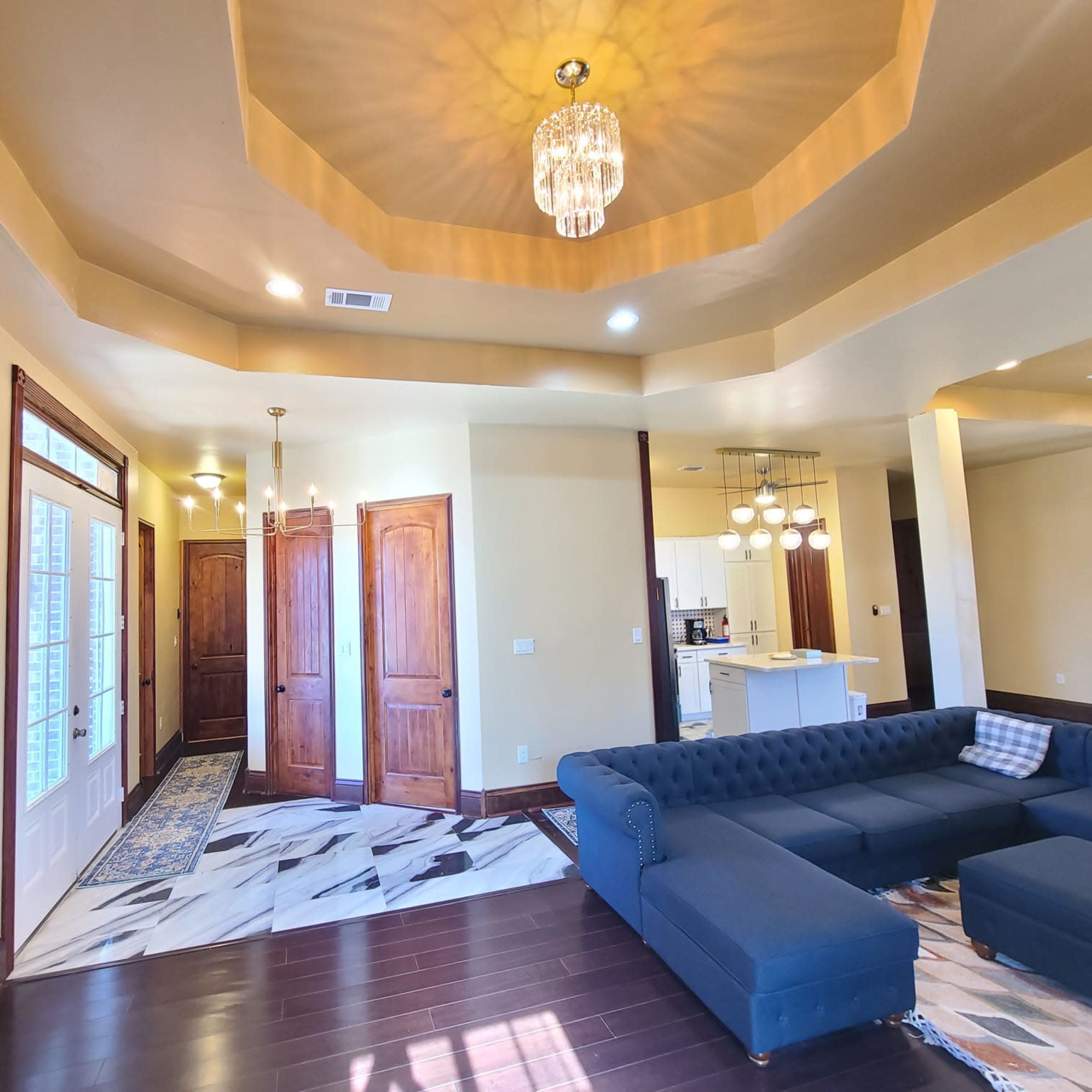 The living room features a blue sectional sofa, a coffered ceiling with a chandelier, and doors leading to a hallway.