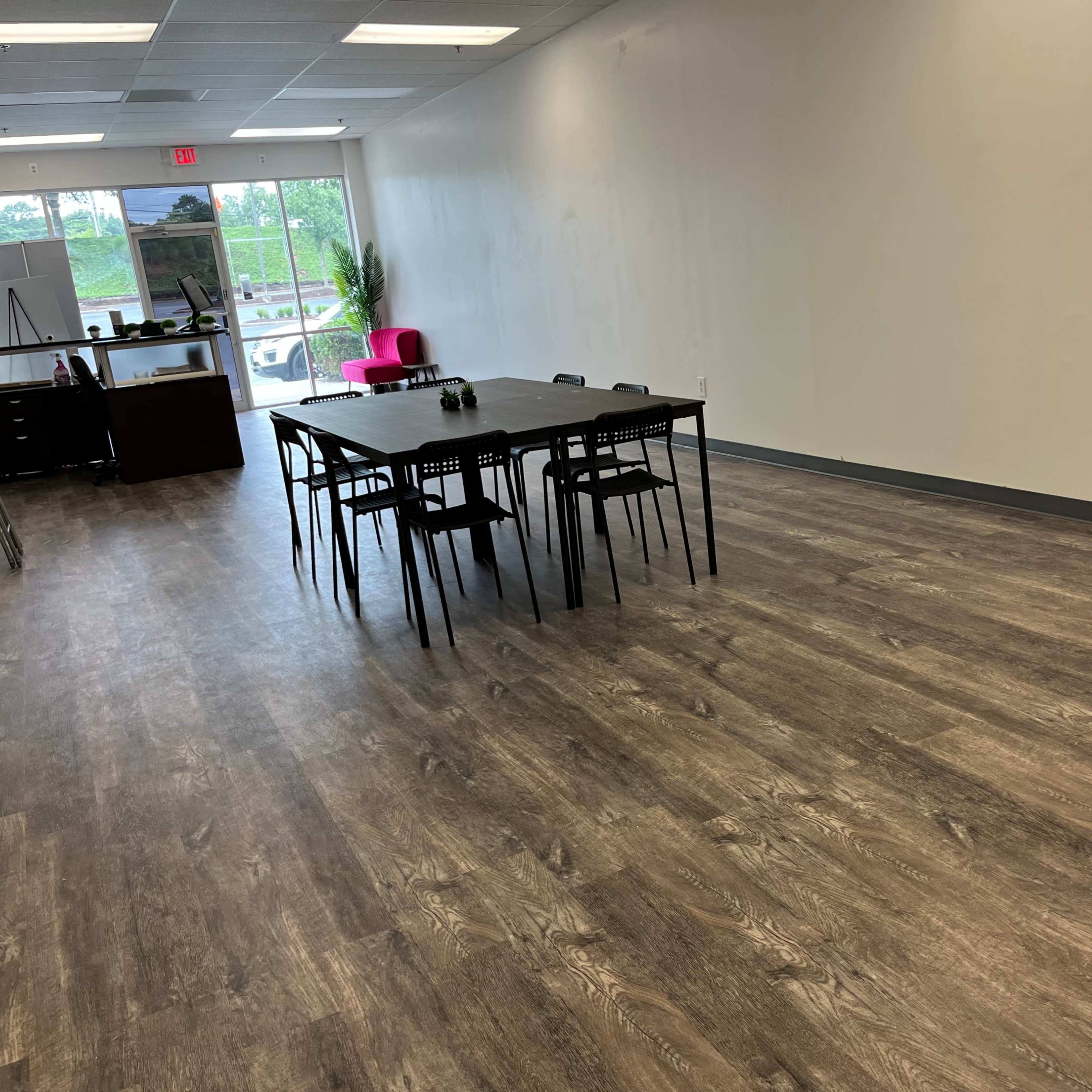 A spacious, empty room with a long black table surrounded by chairs, a small desk in the background, and large windows letting in natural light.