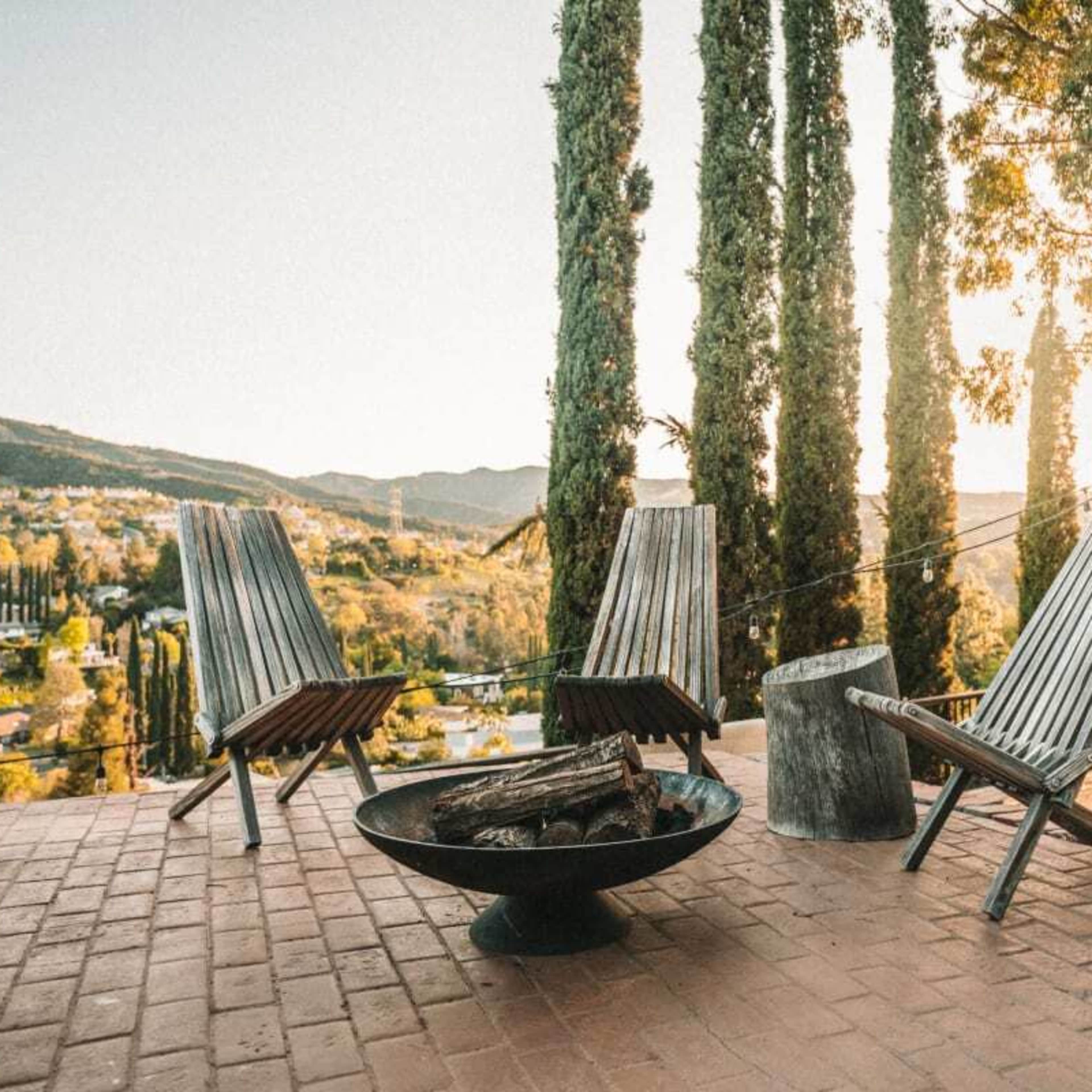 A cozy outdoor seating area features three wooden lounge chairs and a fire pit, set against a backdrop of tall trees and rolling hills at sunset.