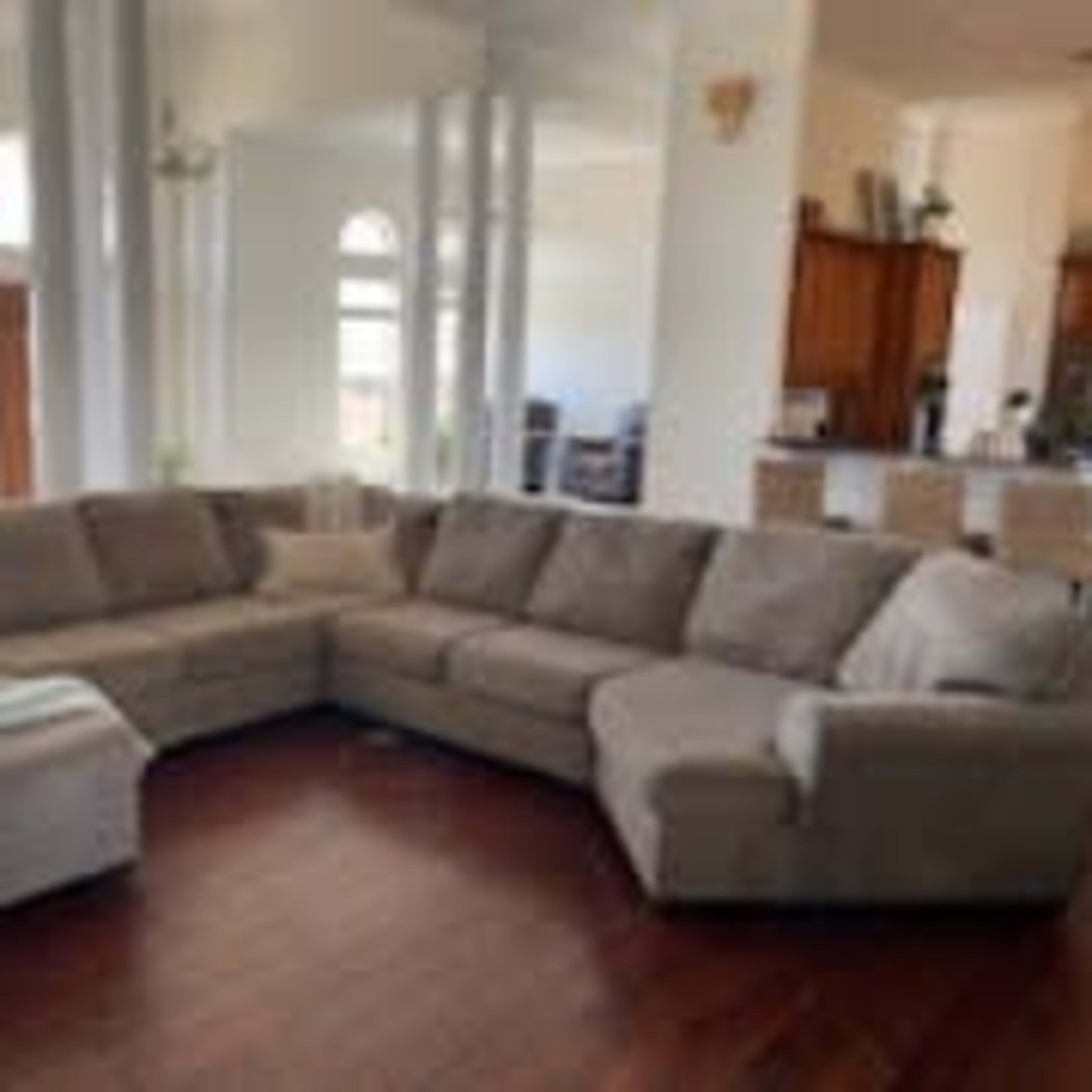 A large, sectional sofa occupies a bright, open living space with wooden flooring and a view of a kitchen in the background.