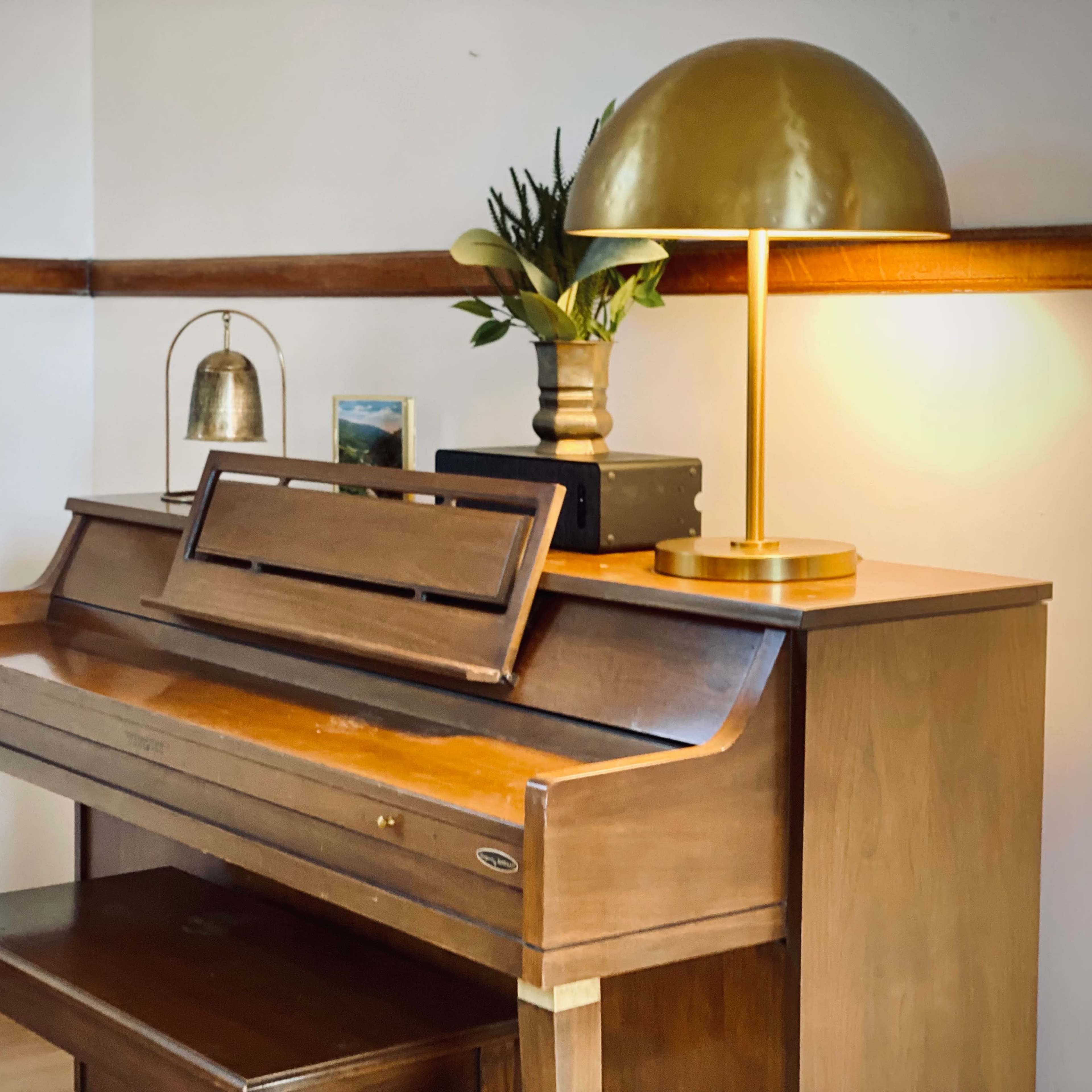 A wooden piano sits against a wall, accompanied by a gold lamp, a small bell, a potted plant, and a framed picture.