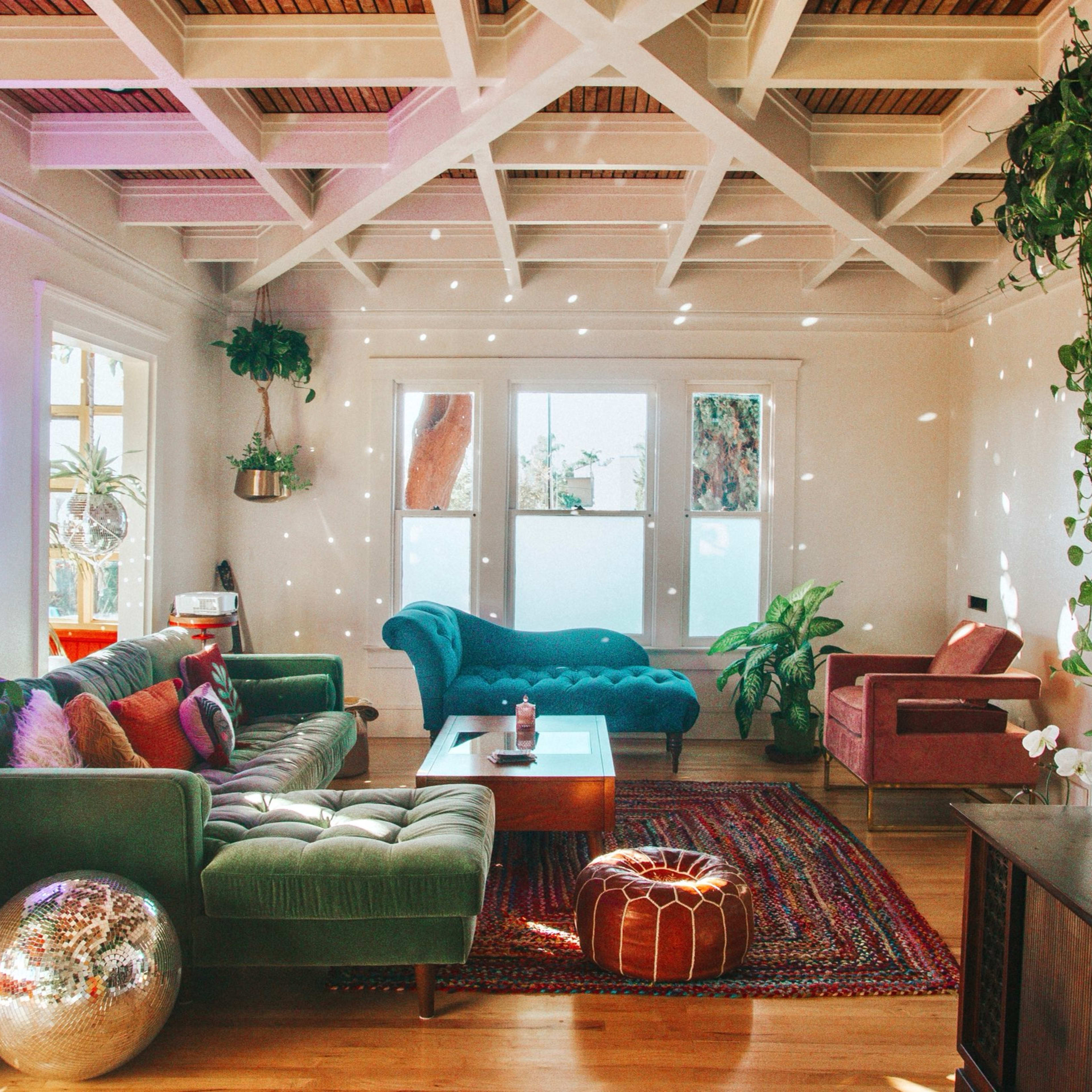 A sunlit living room with a green sofa, a pink chair, a wooden coffee table, plants, and colorful decor on the walls.