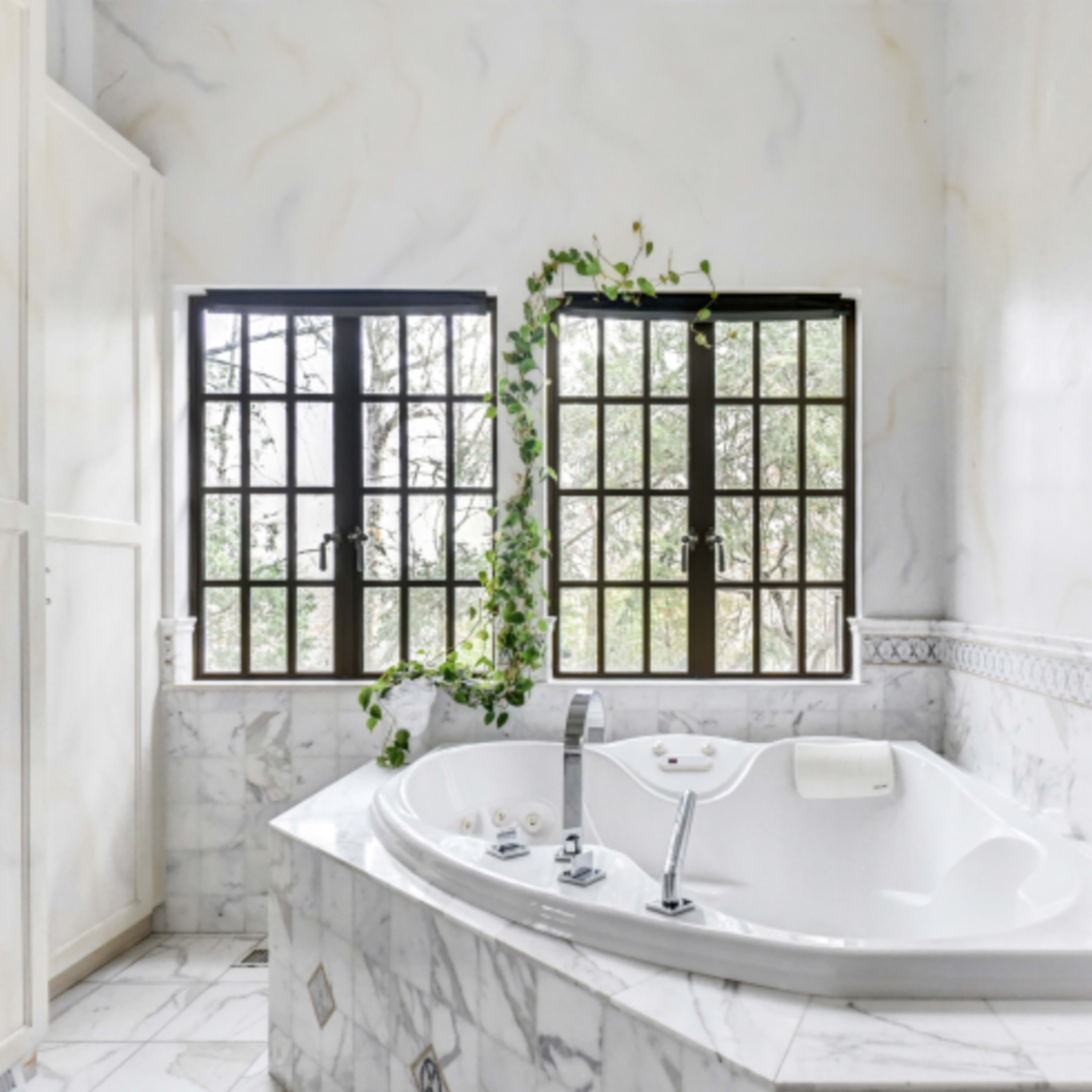 A modern bathroom with a large, white jetted tub surrounded by marble walls and two tall, paneled windows with greenery visible outside.