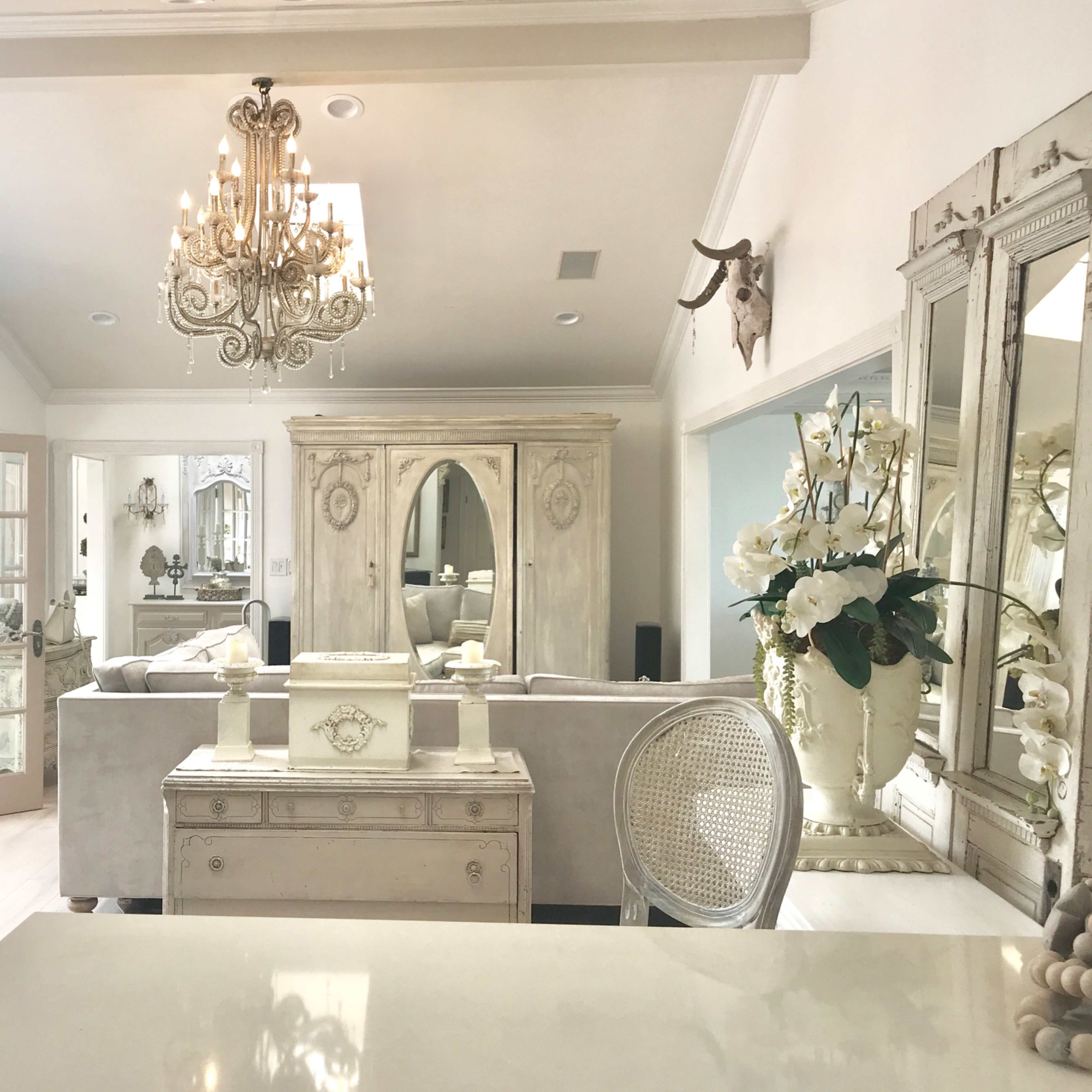 A light-colored living room with a chandelier, a piano, and elegant furnishings, including a large mirrored cabinet and decorative floral arrangements.