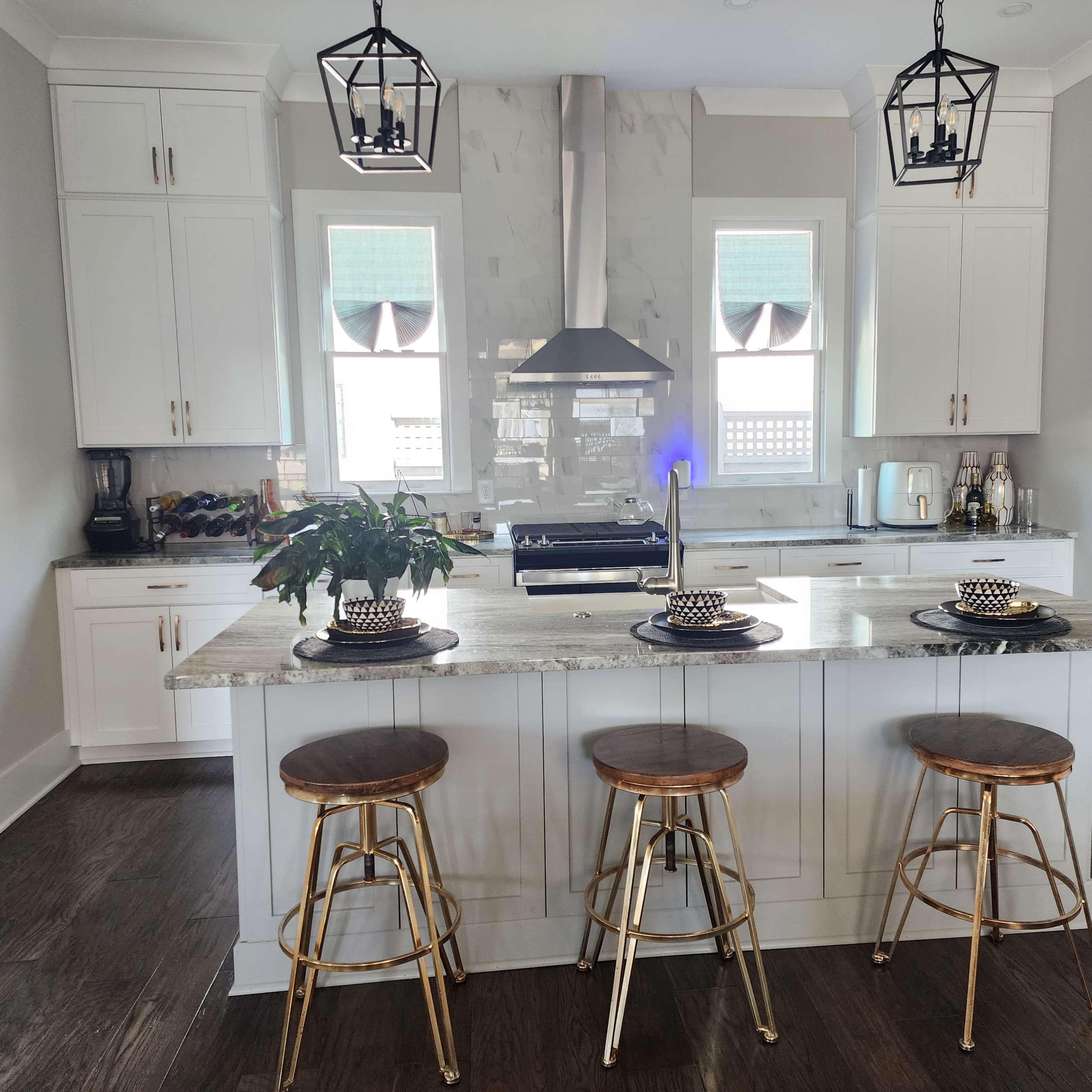 A modern kitchen features a central island with three metal bar stools, pendant lighting, and stainless steel appliances.