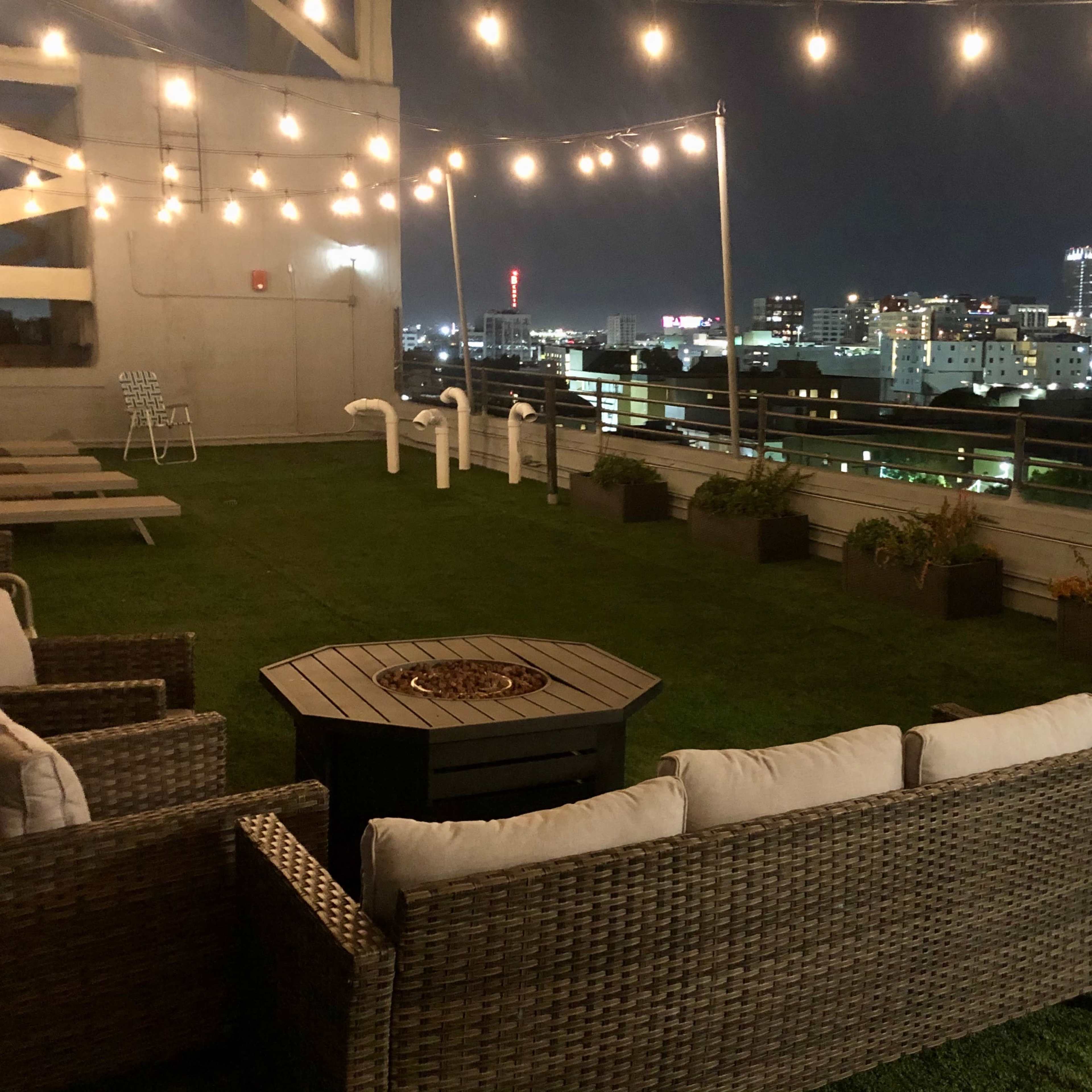 A rooftop terrace with outdoor seating, a fire pit, and string lights, overlooking a city skyline at night.