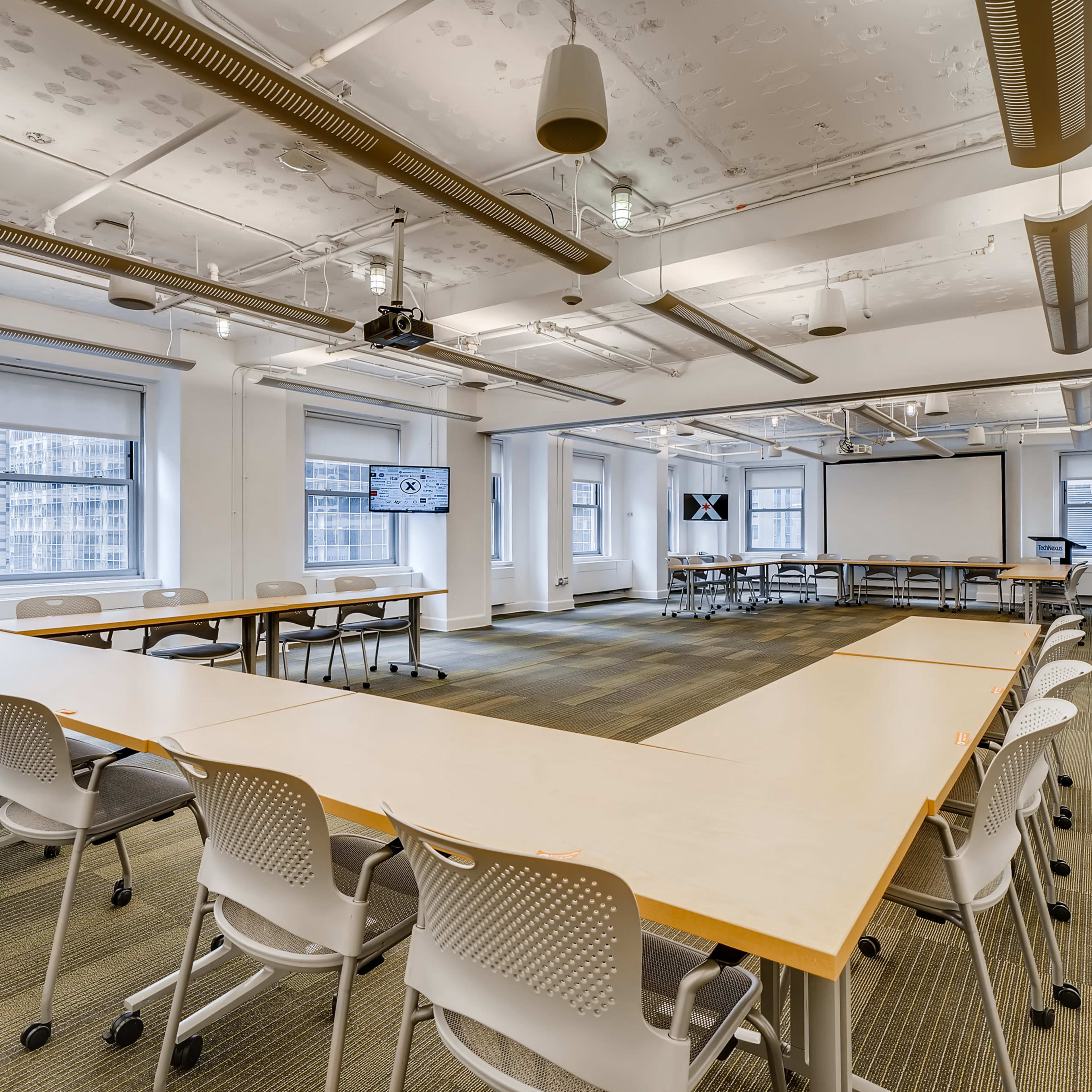 A spacious conference room with multiple tables arranged in a U-shape and chairs surrounding them, with large windows providing natural light.