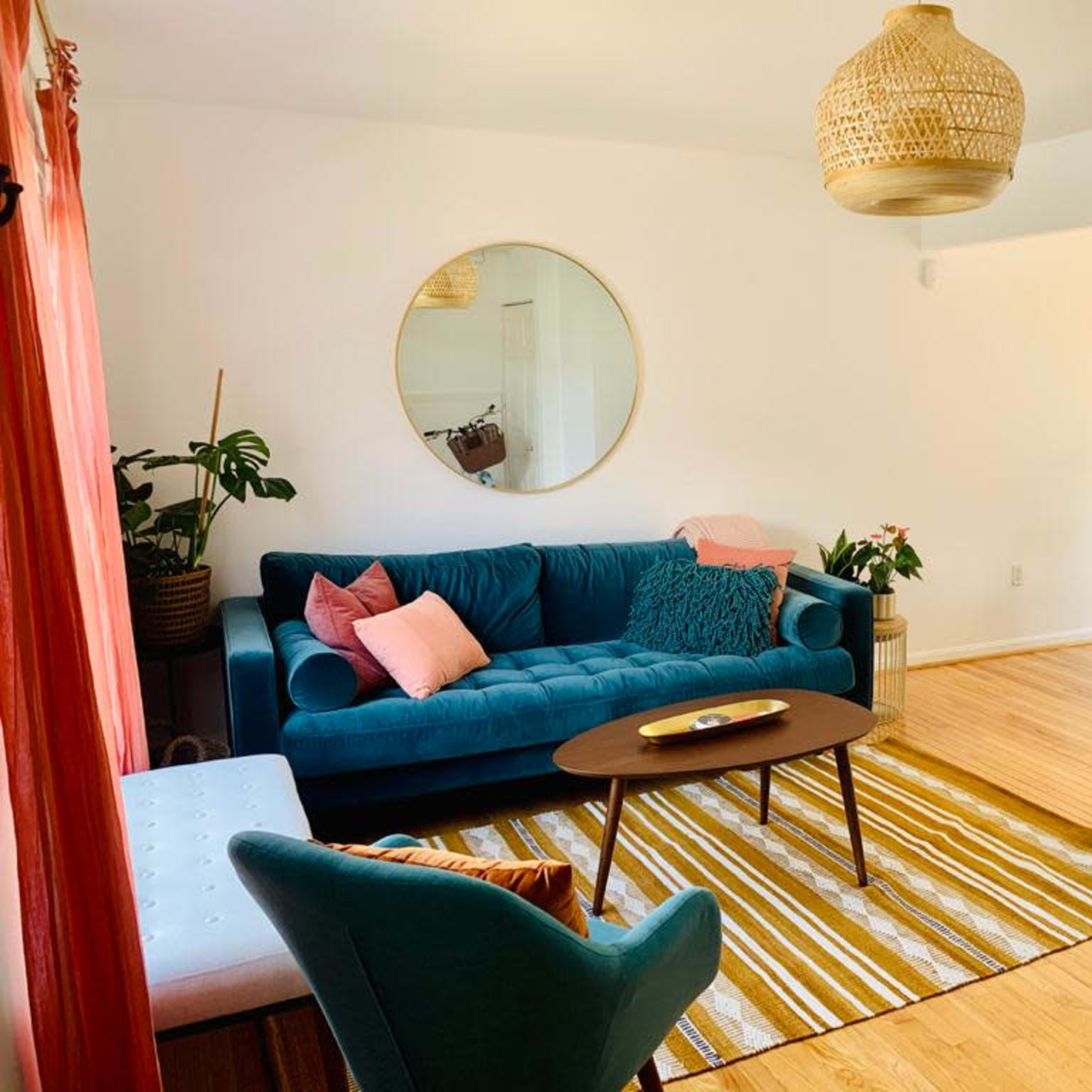 A cozy living room featuring a teal sofa, two teal armchairs, a round mirror, and a wooden coffee table on a striped rug.
