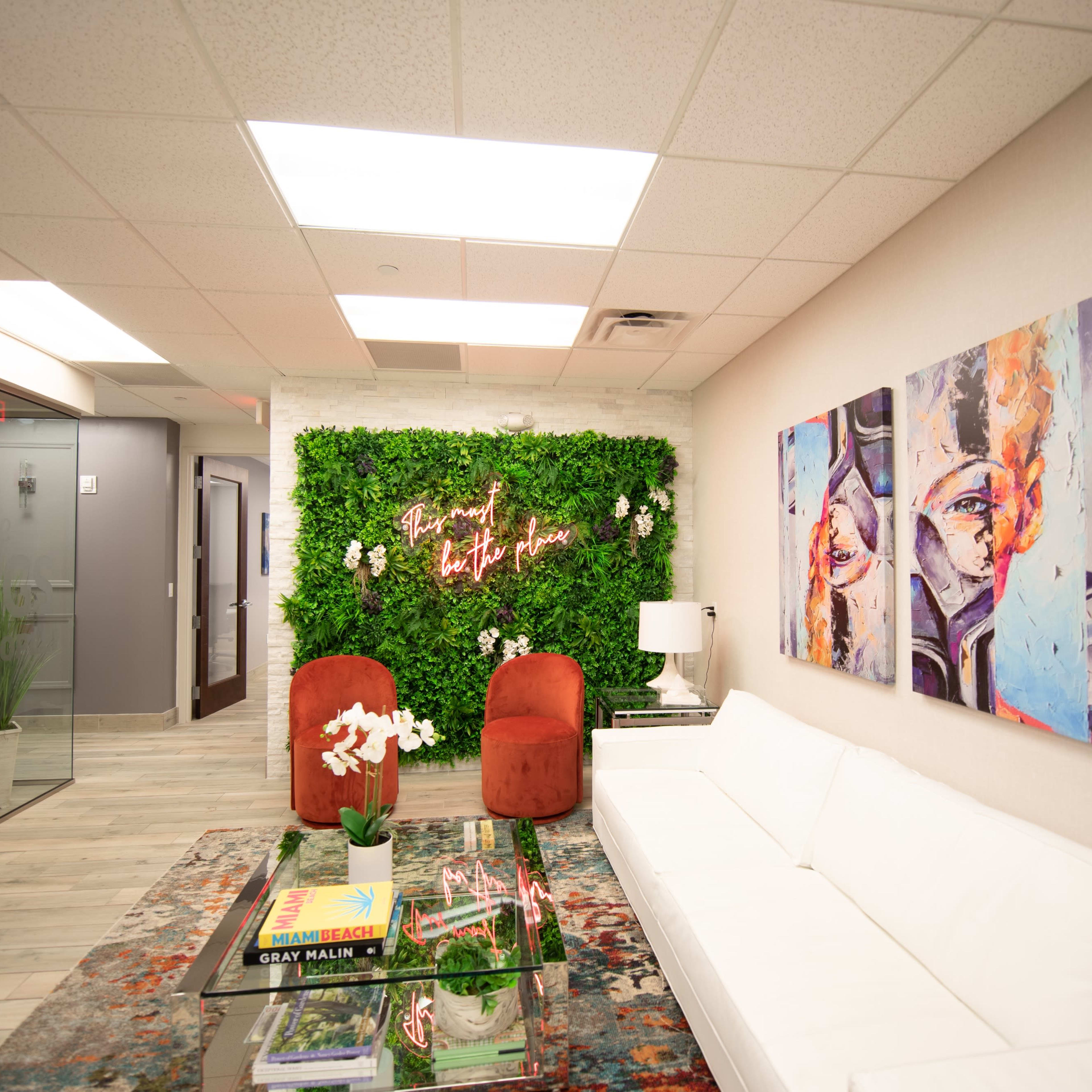 A modern office reception area featuring a white sofa, two red chairs, colorful artwork on the walls, and a green plant wall accent.