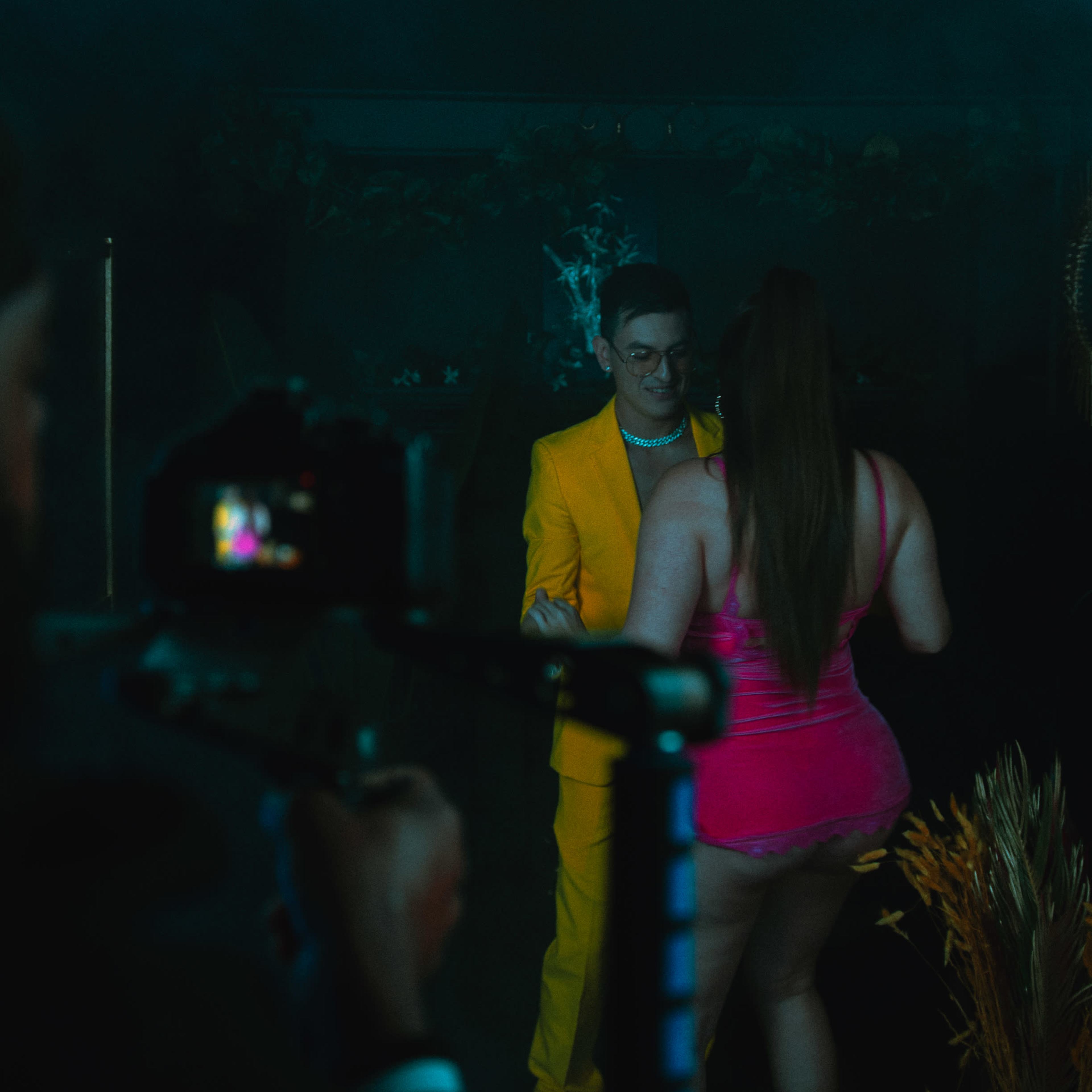 A man in a bright yellow suit dances with a woman in a pink outfit in a dimly lit room filled with greenery.