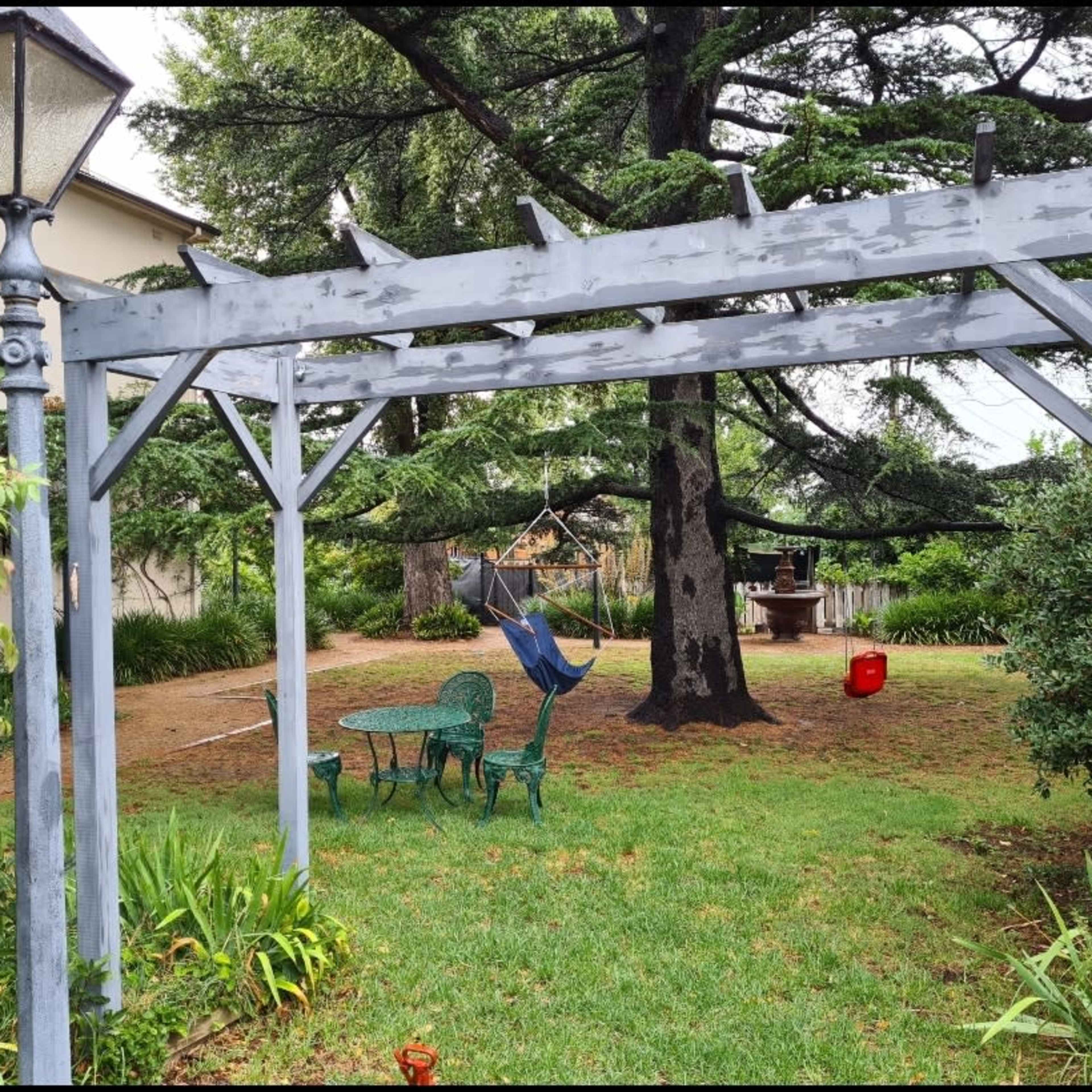 A wooden pergola with a lamp post frames a small seating area featuring a green table and chairs in a grassy garden.