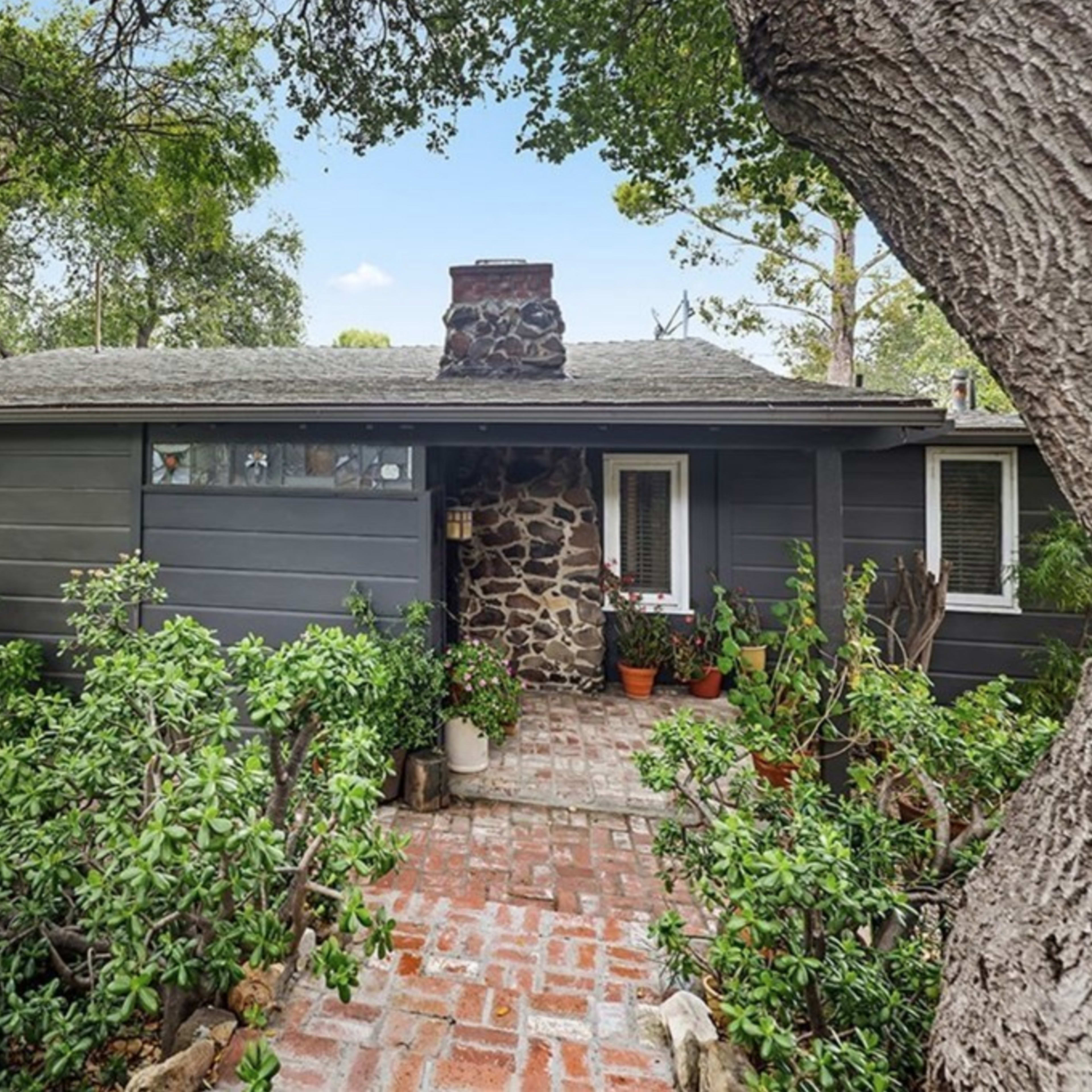 A single-story house with a stone chimney is surrounded by greenery and a brick pathway leading to the entrance.