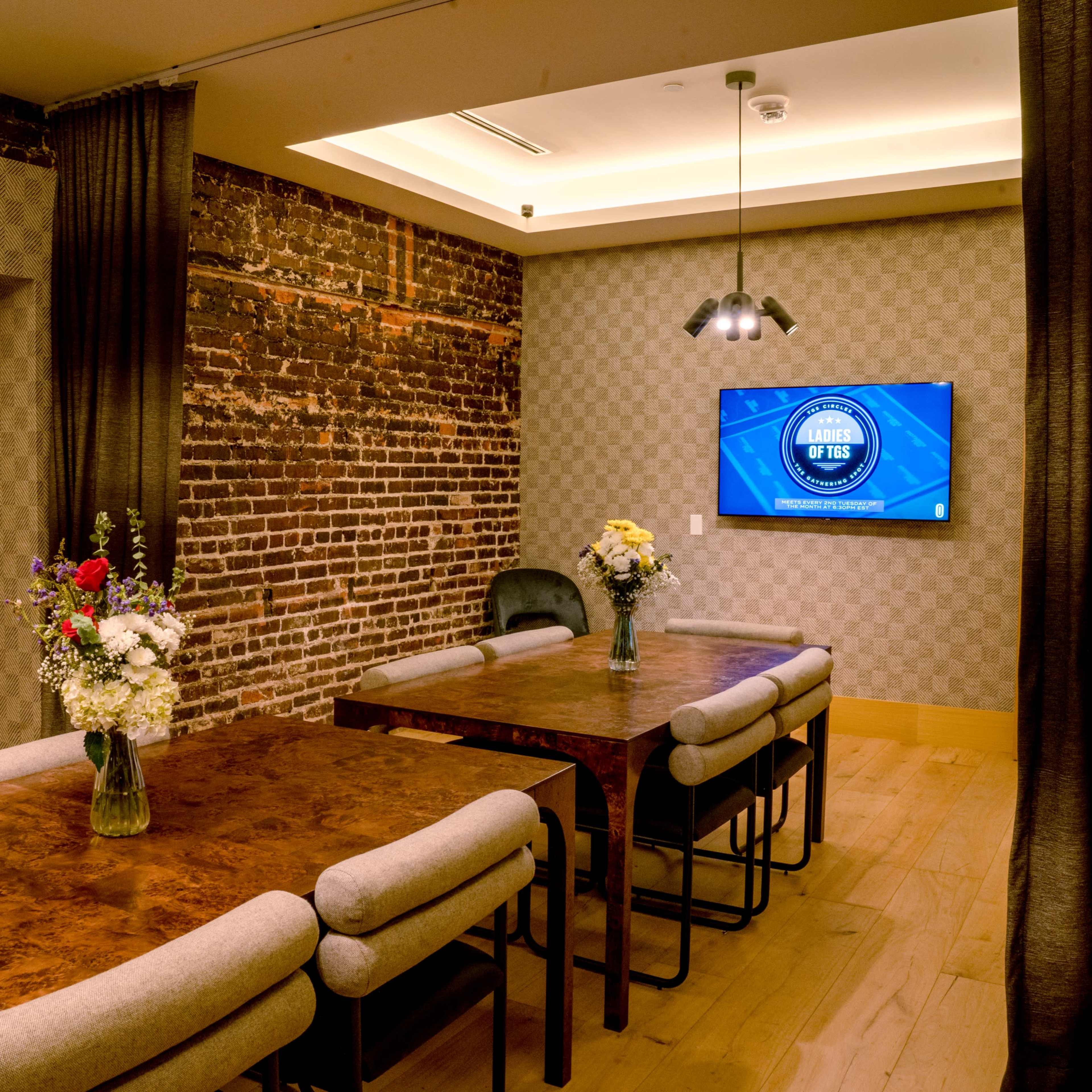 A conference room features a long wooden table surrounded by padded chairs, with a large screen mounted on the wall and floral centerpieces on the table.