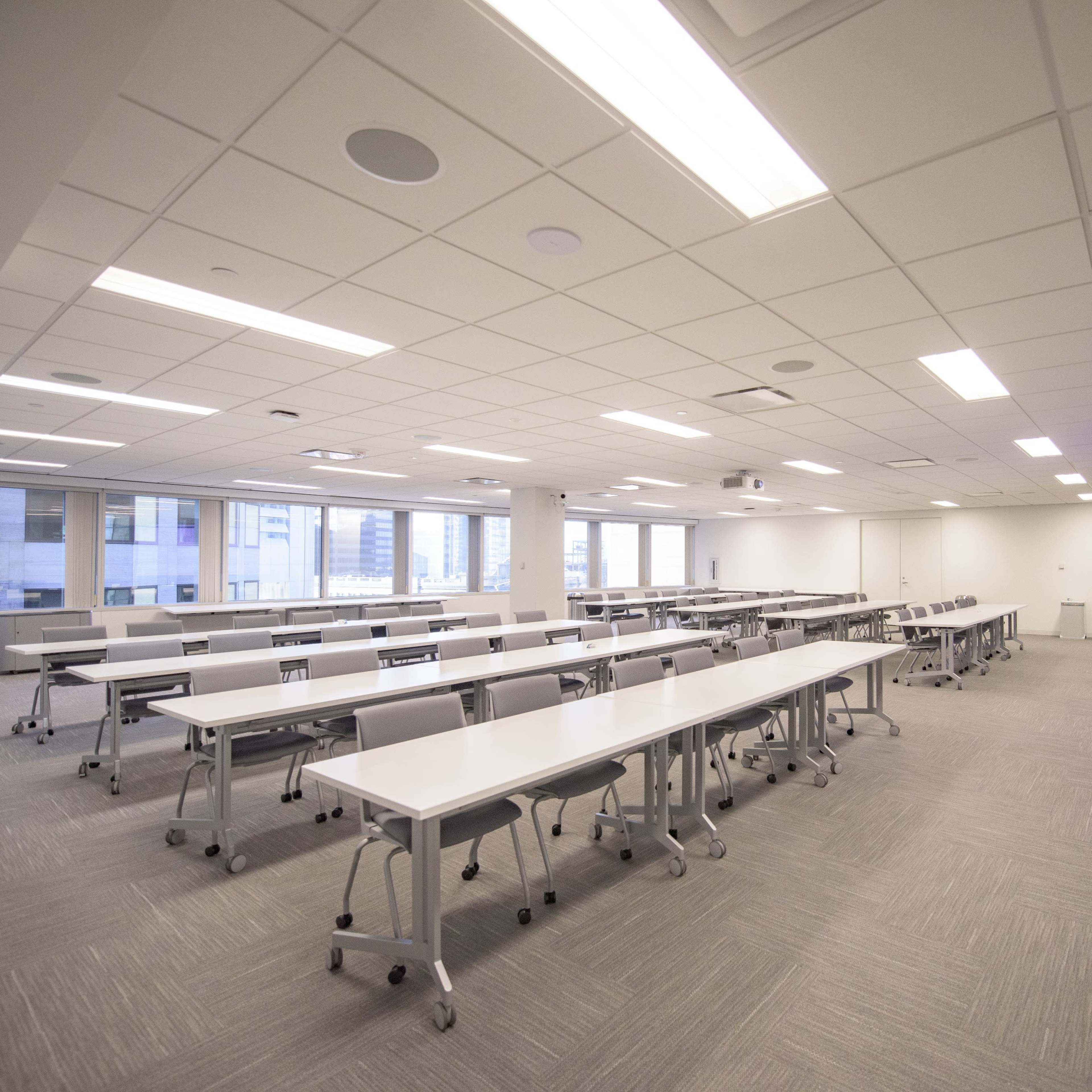 A spacious conference room features multiple tables arranged in rows, with a large screen at one end and windows providing natural light.