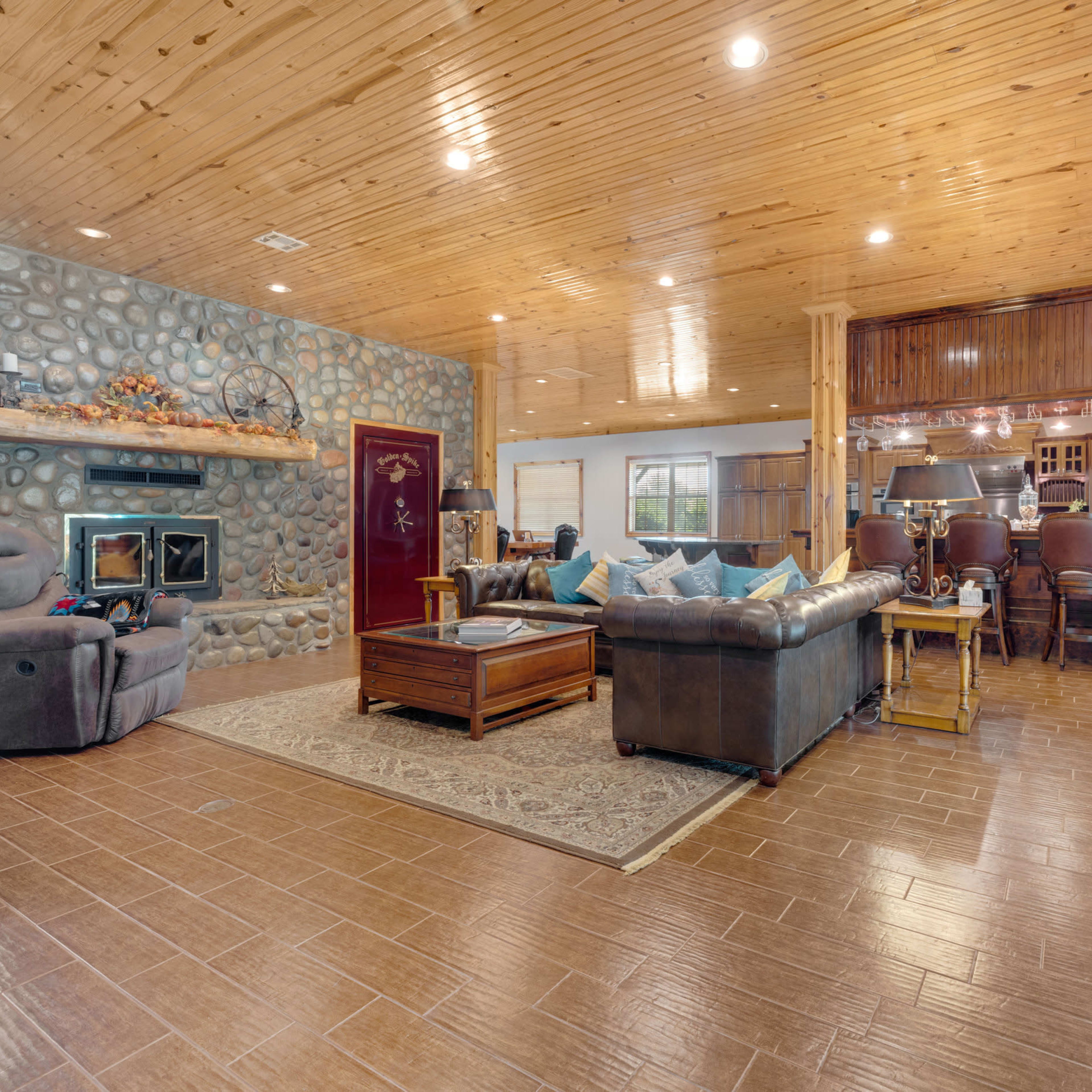 A spacious living room with a stone fireplace, wooden ceiling, and a seating area arranged around a coffee table, adjacent to a kitchen with a bar.