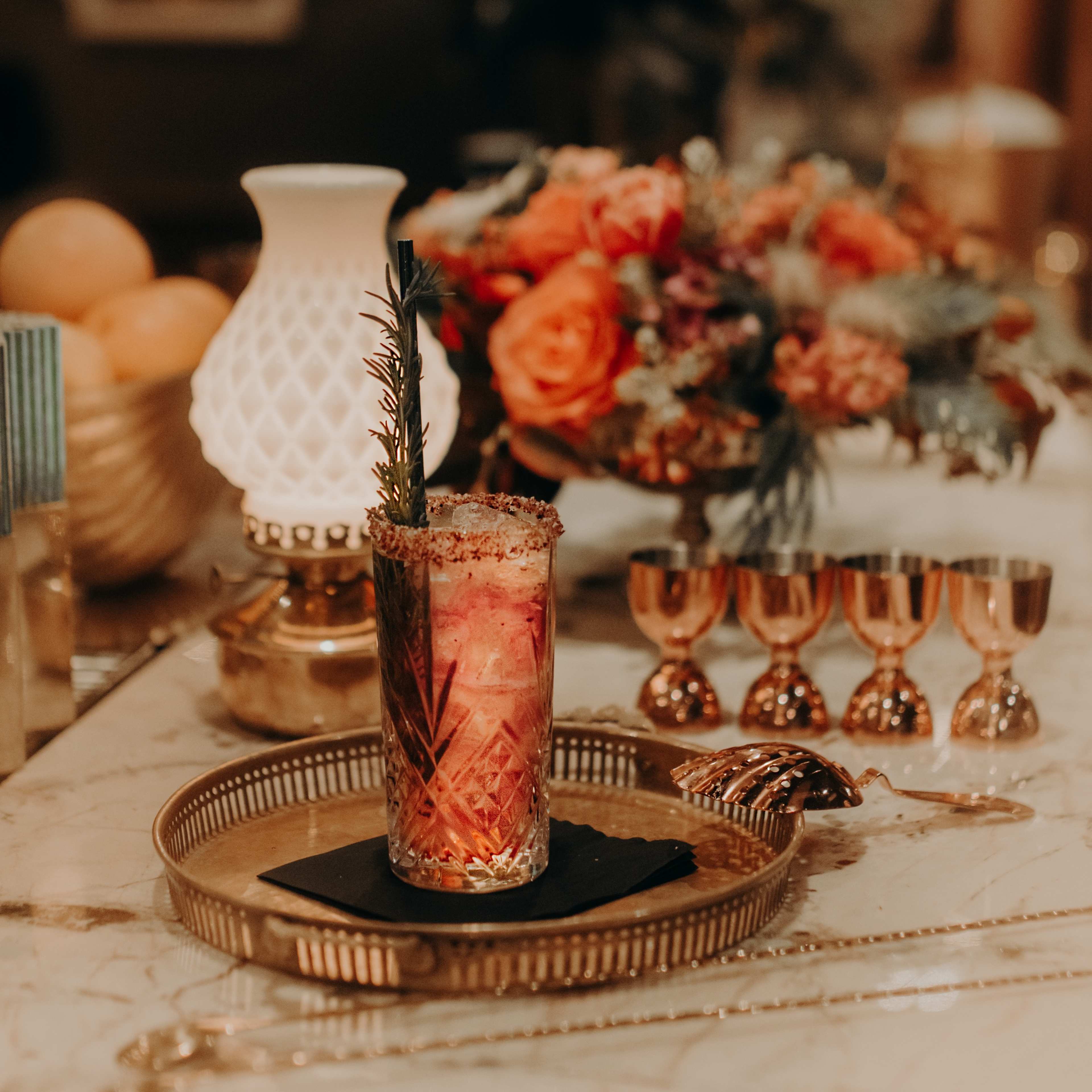 A cocktail garnished with a sprig of rosemary is presented on a decorative tray alongside a bouquet of flowers and several small metal cups.