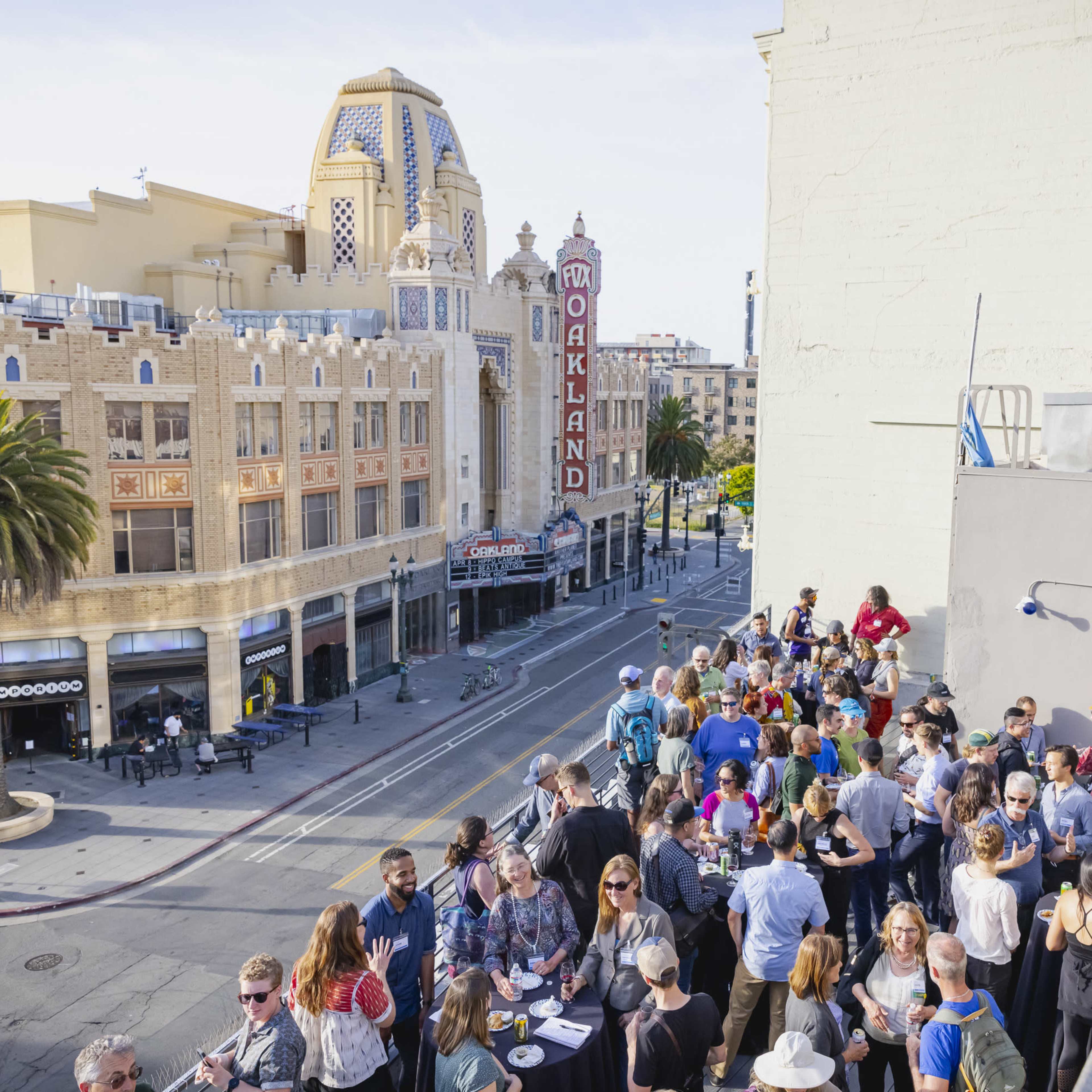 A large group of people gathers on a rooftop overlooking a street in Oakland, with an art deco theater visible in the background.