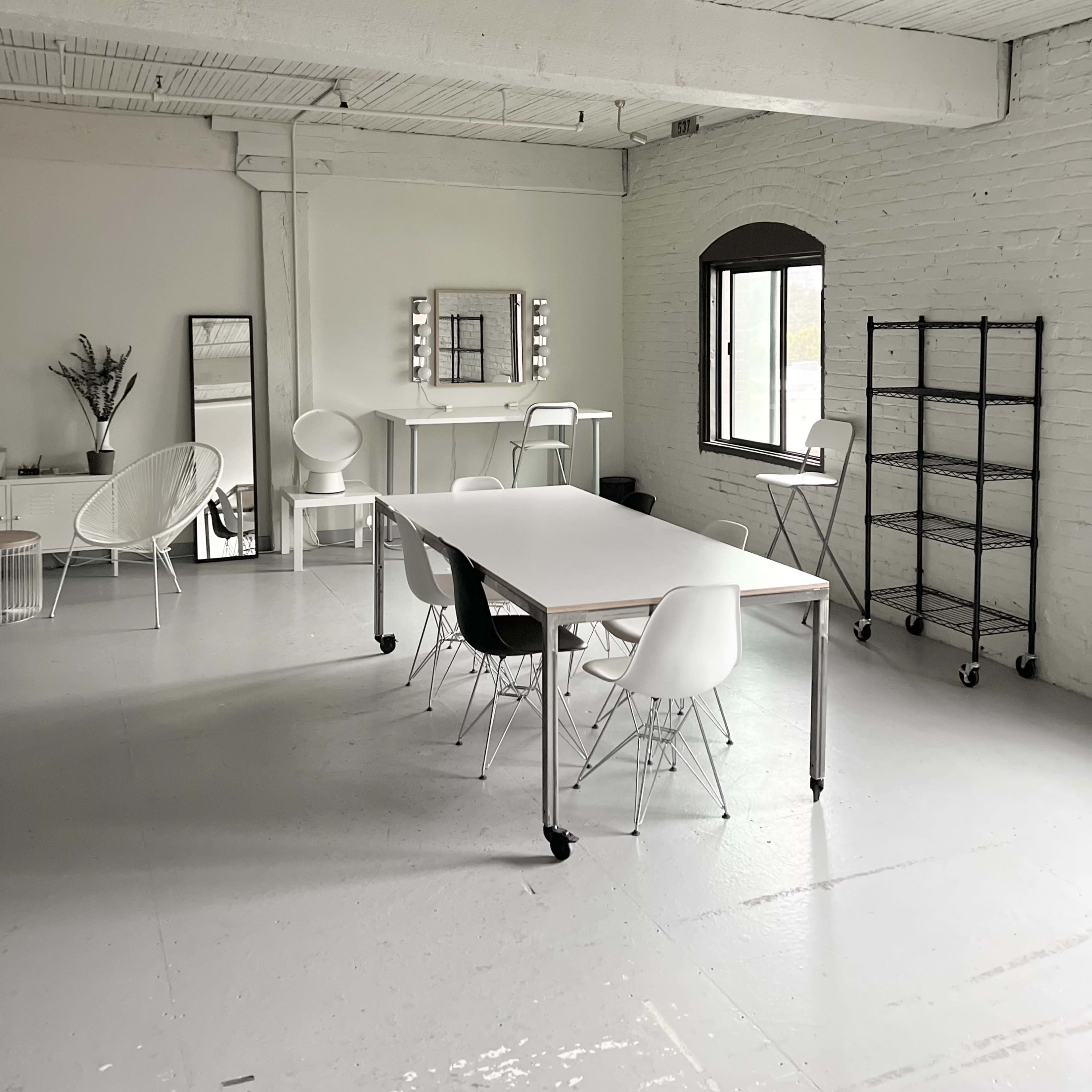 A spacious, well-lit room featuring a long table surrounded by chairs, a mirror on the wall, and a shelving unit.
