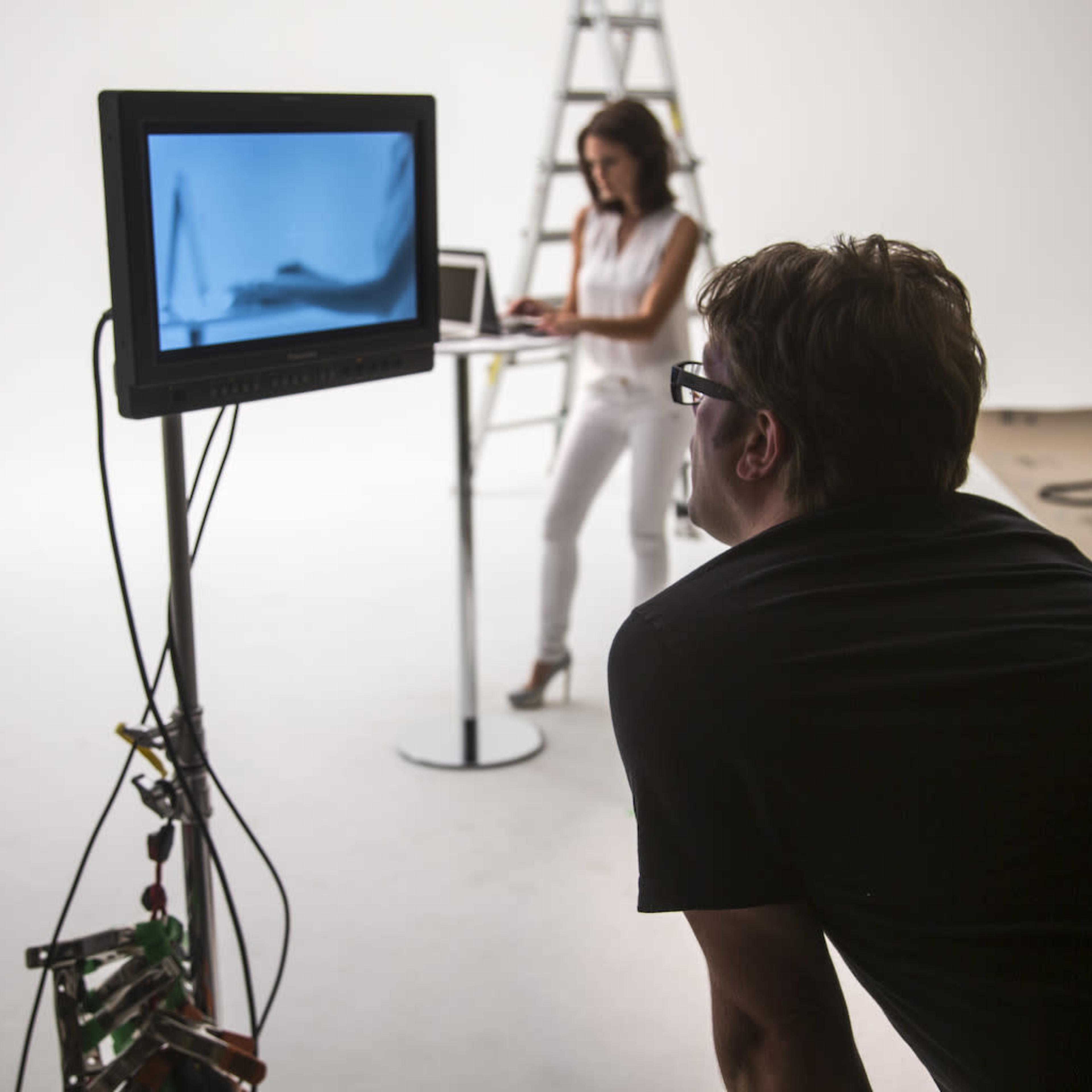 A man observes a monitor displaying live footage while a woman works on a laptop in a photography studio.
