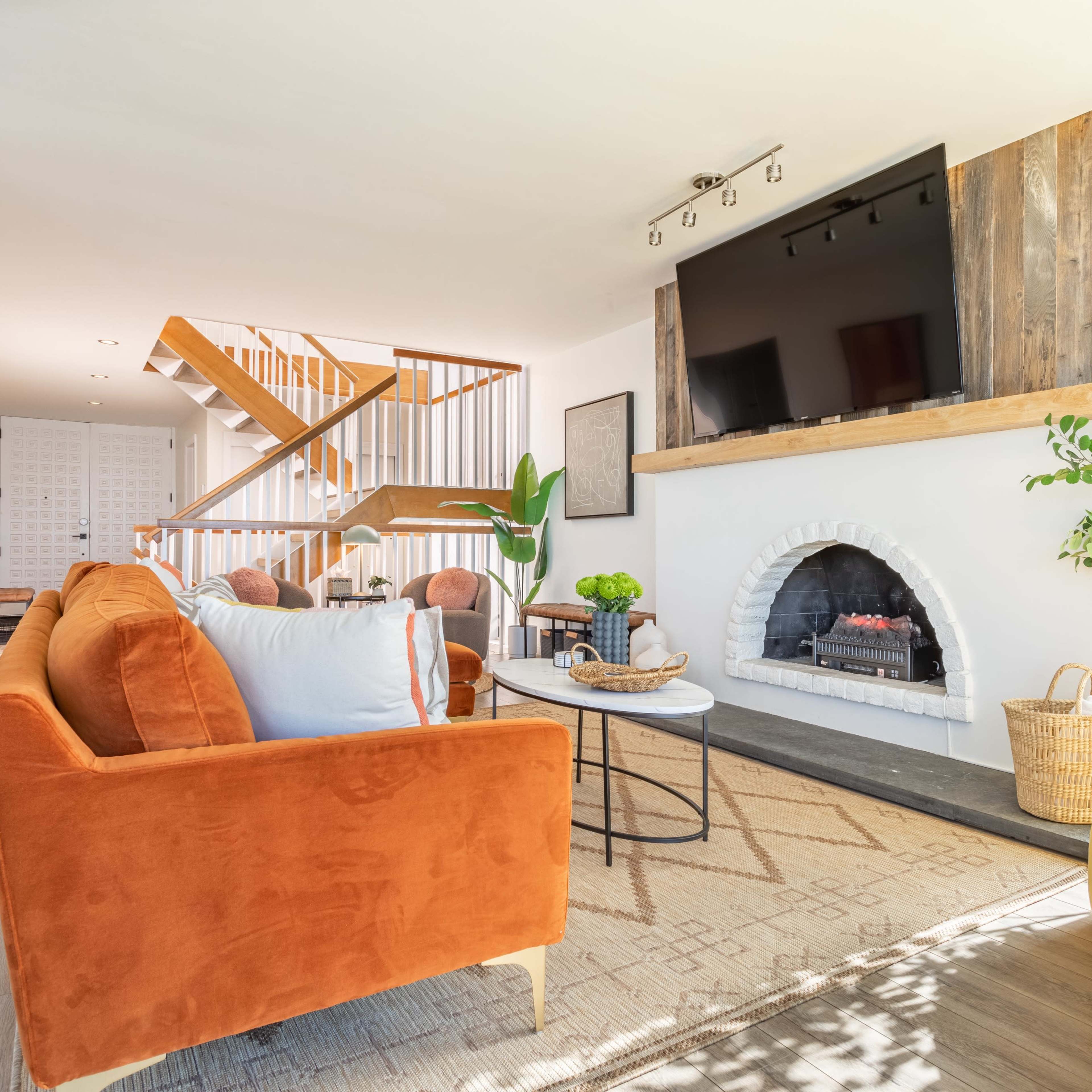 A modern living room featuring an orange couch, a round coffee table, a white fireplace, and a staircase in the background.