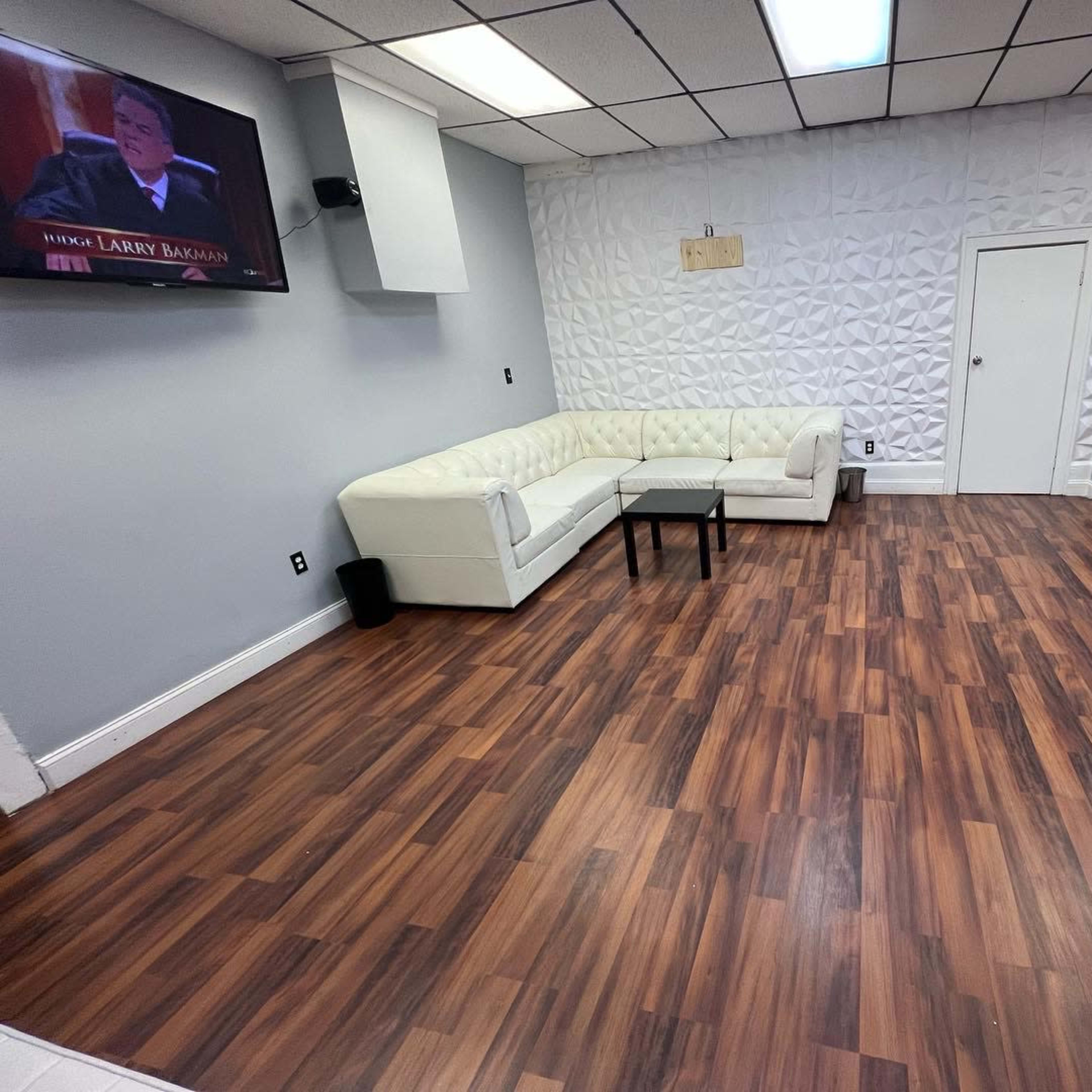 A sparsely furnished room with a white sectional couch, a television mounted on the wall, and a wooden floor.