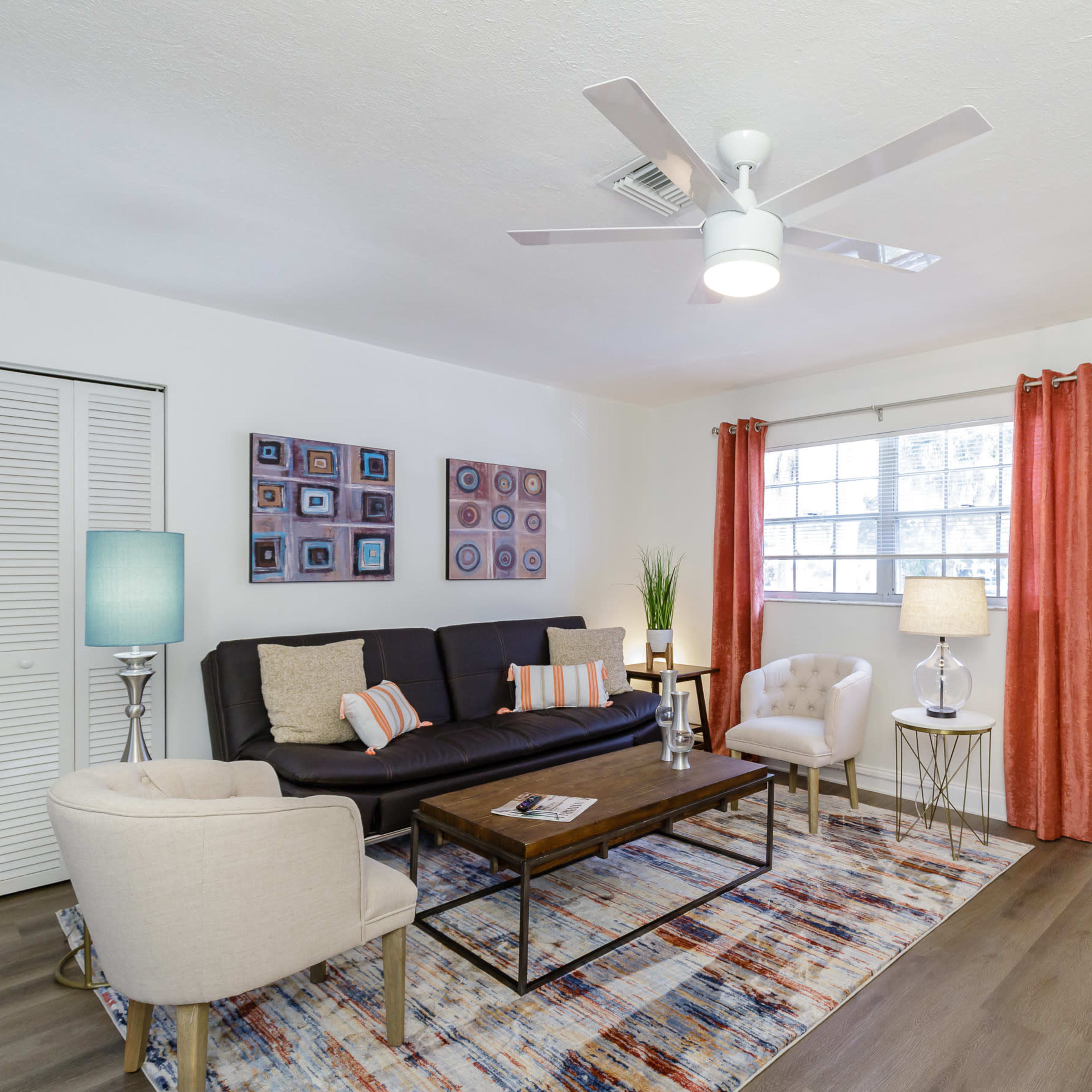 A living room with a black sofa, beige armchair, wooden coffee table, and a large window draped with orange curtains.